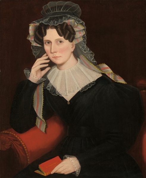 The image depicts a woman seated in a three-quarter view. She has light skin, delicate facial features with slightly arched eyebrows, and her hair is styled with prominent coils. She is holding a small red book, with her left hand resting against her face. She wears a dark gown with long sleeves, a lace collar, and lace cuffs. She also wears a black cap trimmed with lace and a ribbon in pink, blue, and yellow. The background is deep brown, and she is seated on a red velvet chair with gold studs along the edge.