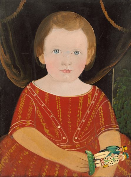 The image shows a portrait of a young child from the waist up, facing forward. The child has light skin, blue eyes, short brown hair, and wears a red outfit with gold and white patterns. They hold a small, colorful object and stand in front of dark curtains with decorative tassels, with a hint of greenery visible in the background.