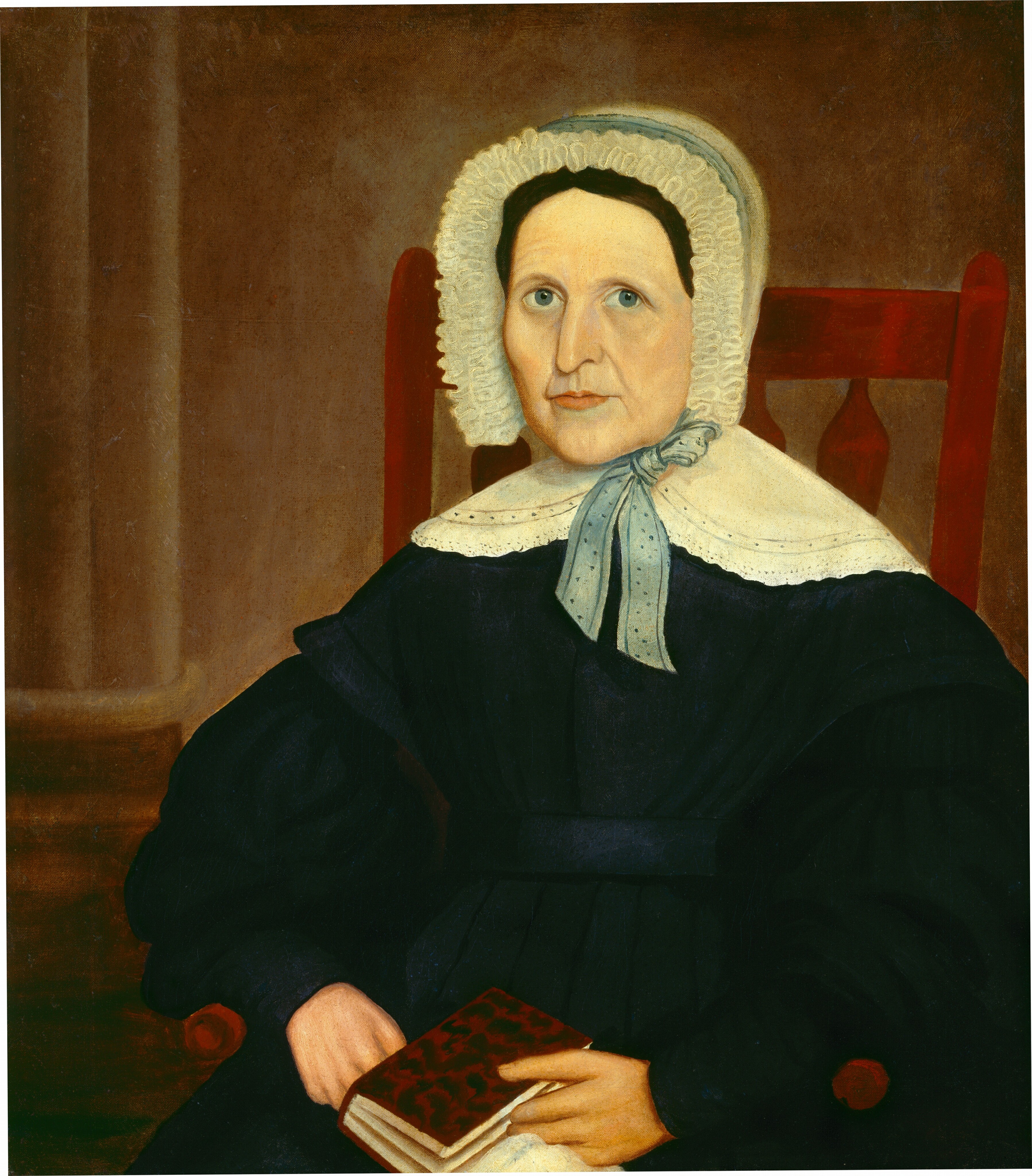 The image shows an older woman depicted from the waist up, seated in an upright position. Her facial features include fair complexion, prominent cheekbones, and blue eyes, with dark hair mostly hidden under a white bonnet with a frilled edge. She wears a dark-colored dress with full sleeves and a white detachable collar tied at the front with a blue ribbon. The woman holds a small book with her left hand resting on the armrest of the chair and the right hand gripping the book. The background includes a plain brownish wall.