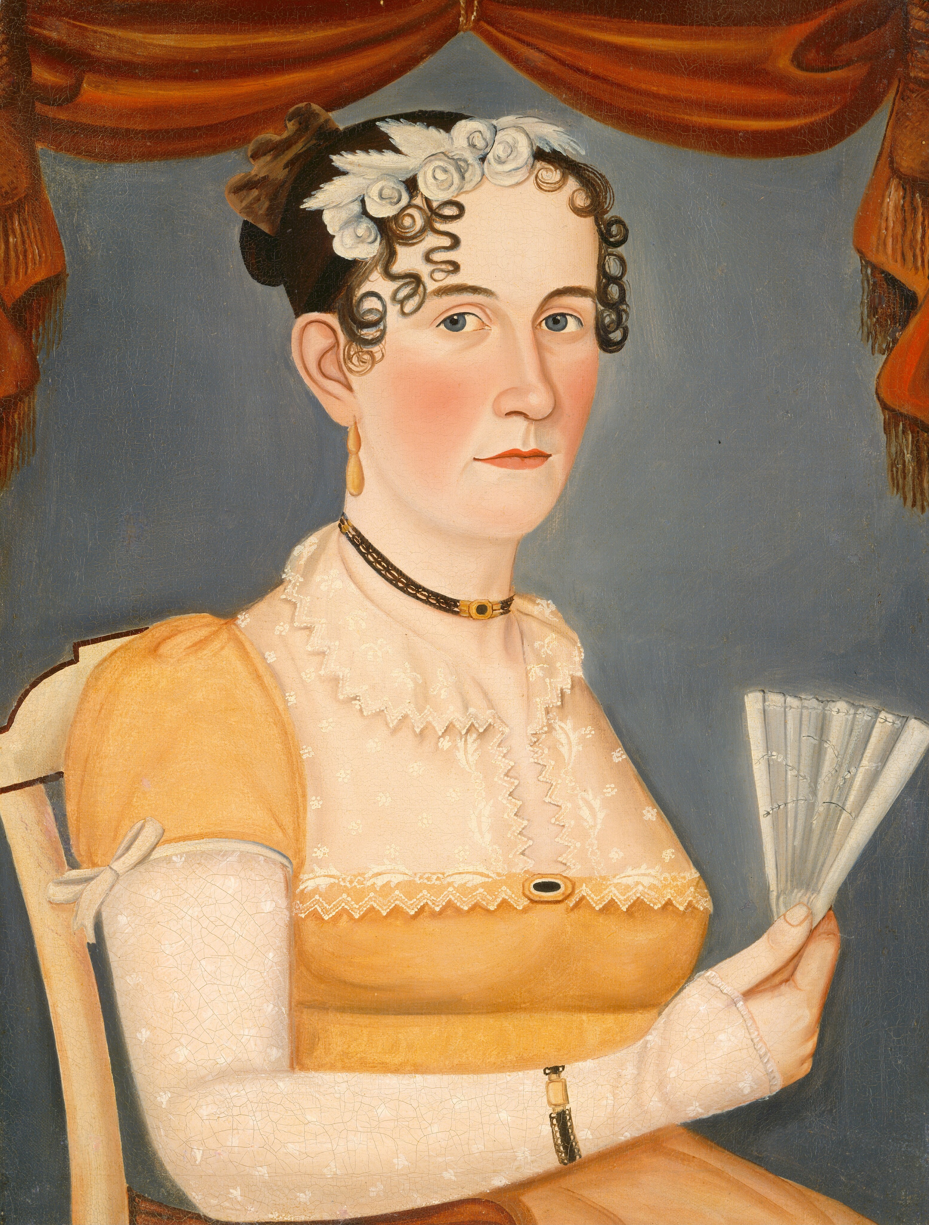 The image depicts a woman's upper body. She is seated in a sideways position but facing forward towards the viewer. The woman has dark hair styled in an updo with ringlet curls framing her face. Her facial features include fair complexion with a slight blush on her cheeks and blue eyes. She wears a yellow dress with puffed sleeves, patterned white lace detailing on the bodice, and a high waistline. Around her neck is a black choker with a gold pendant, and she has delicate gold drop earrings. She wears a bracelet on her right wrist and holds an ornate white fan in her right hand. The background is a plain, muted blue, with a reddish-brown drapery with tassels framing the top and sides of the portrait.