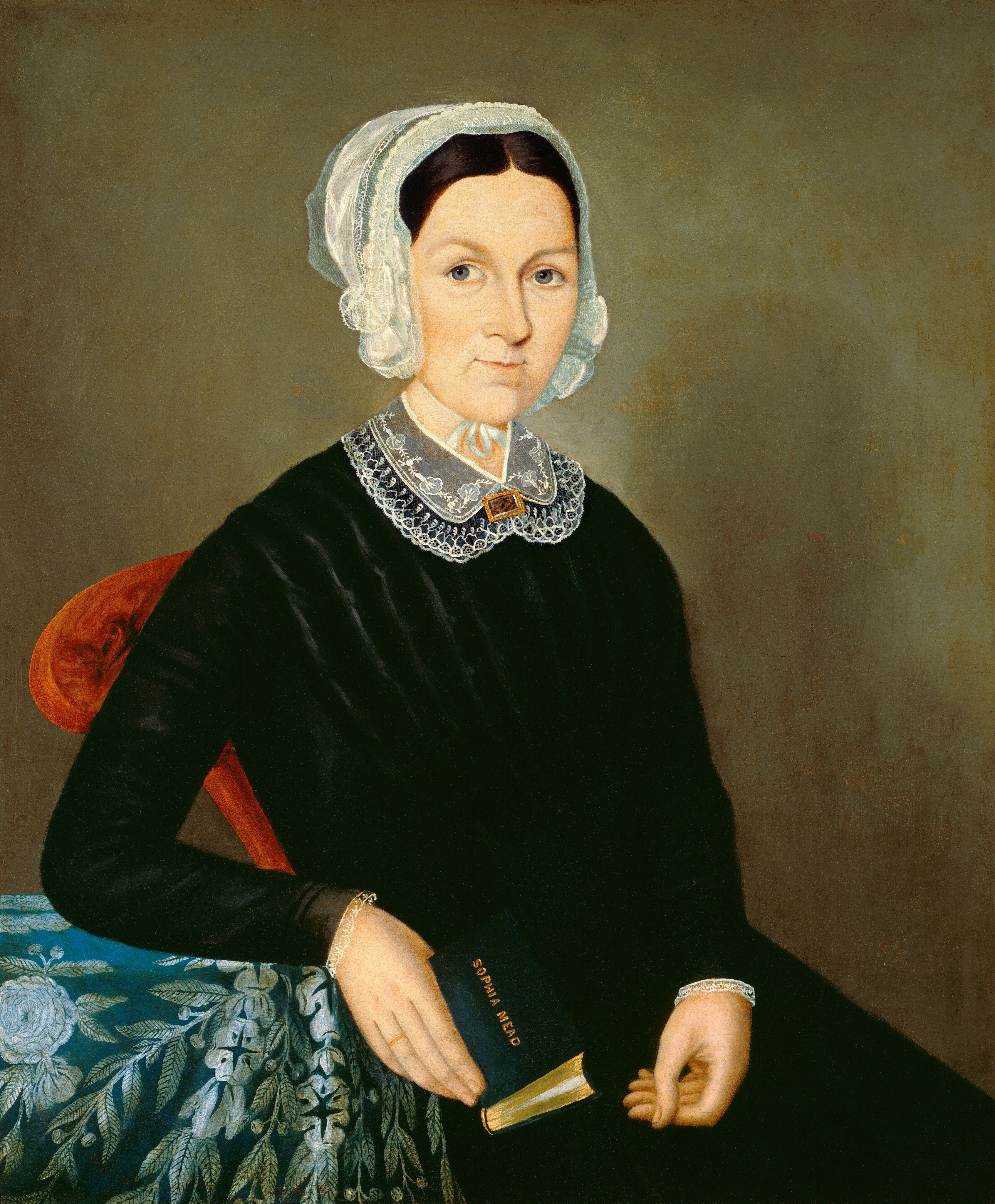 The image shows a portrait of a woman seated in a chair. She is facing slightly to the left and looking directly at the viewer. She has fair skin, light-colored eyes, and dark brown hair styled under a lace-edged cap. She wears a black dress with a lace collar and holds a small book titled "Sacred Songs" in one hand. She also wears a bracelet on her wrist. She is seated in a chair with a red upholstered back and an armrest draped with embroidered blue and green fabric. The background is a plain, muted brown color.
