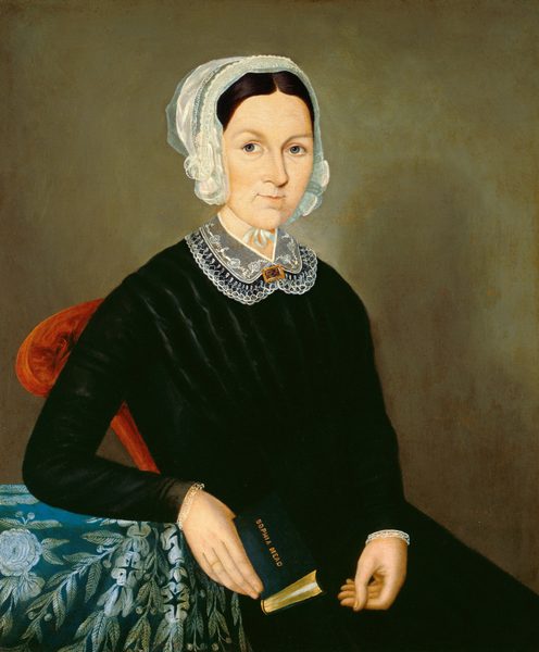 The image shows a portrait of a woman seated in a chair. She is facing slightly to the left and looking directly at the viewer. She has fair skin, light-colored eyes, and dark brown hair styled under a lace-edged cap. She wears a black dress with a lace collar and holds a small book titled "Sacred Songs" in one hand. She also wears a bracelet on her wrist. She is seated in a chair with a red upholstered back and an armrest draped with embroidered blue and green fabric. The background is a plain, muted brown color.