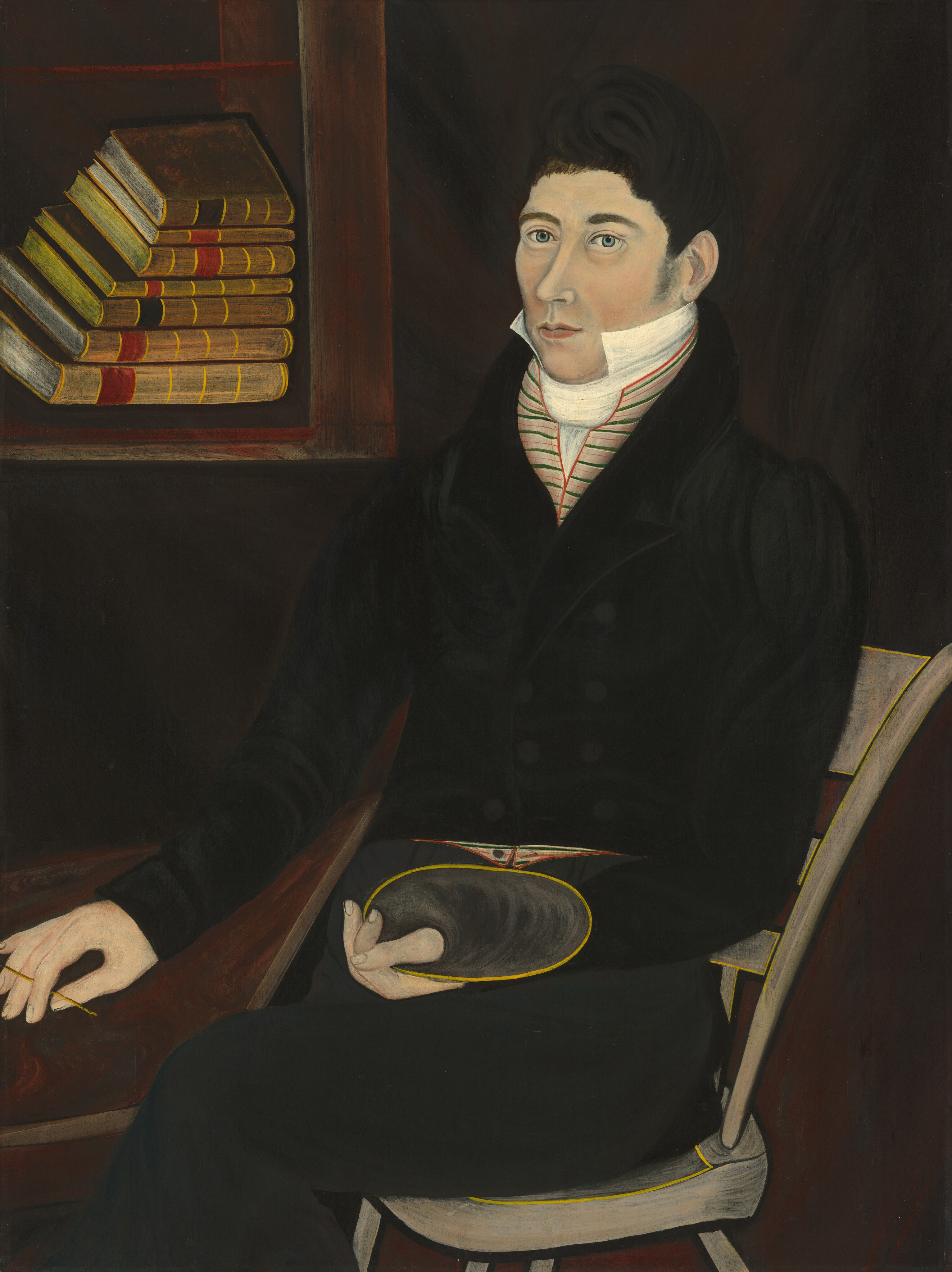 In the image, a man is seated in a wooden chair, facing forward. He has light skin, blue eyes, dark brown curly hair, and wears a high-collared coat over a striped cravat. There is a white cloth around his neck, and he holds a thin object in one hand while a hat rests on his lap. A wooden bookshelf with several books is in the background.