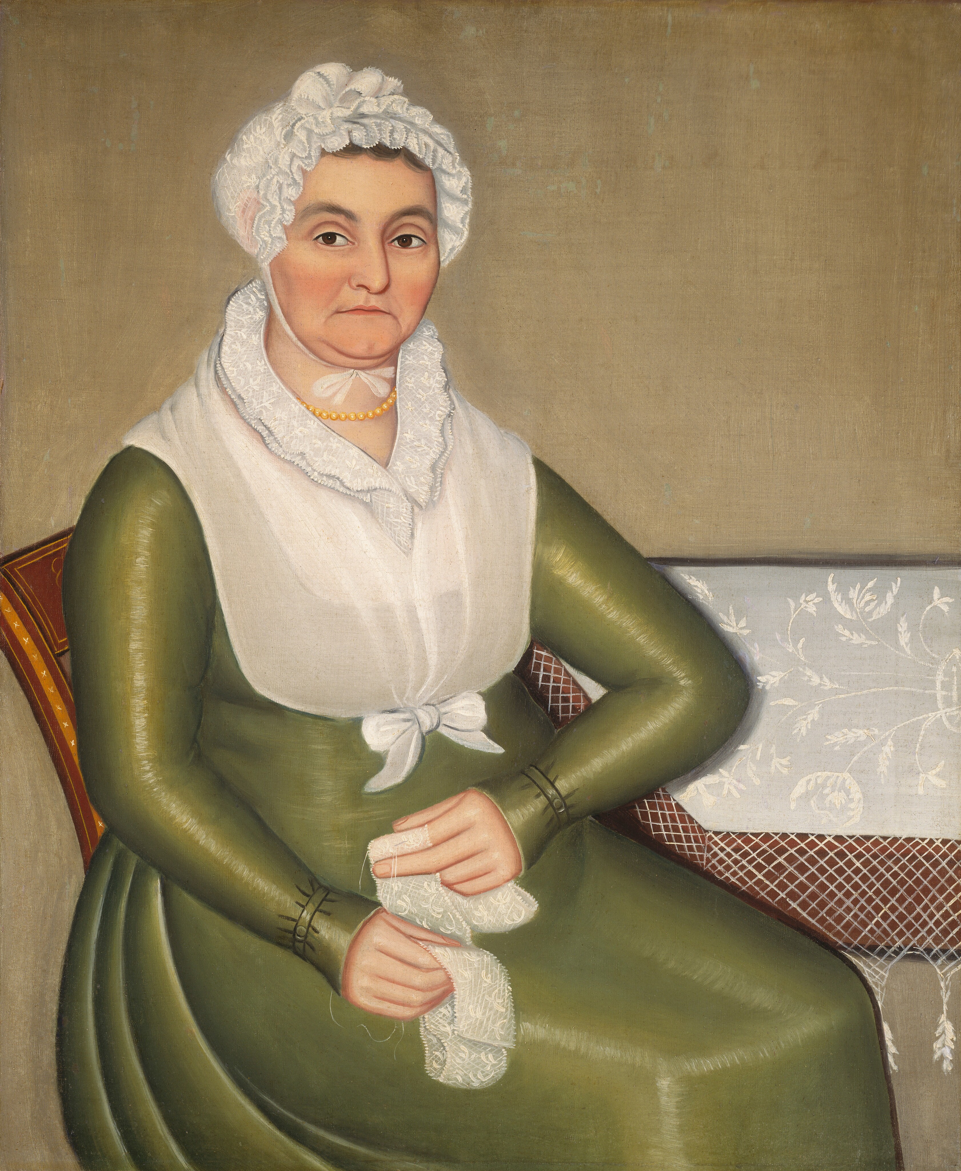 The image depicts a woman from the waist up, seated and holding a piece of lacework. She faces slightly to her right, with a direct gaze. Her facial features include deep-set eyes, a pronounced nose, and slightly pursed lips. She wears a white cap with lace edges and an olive-green dress with long sleeves. A white shawl drapes over her dress and is tied into a bow at the front. She wears a string of pearls around her neck. Beside her is a table with a decorative cloth featuring floral motifs. The background is a plain, muted tone.