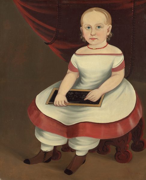 The image depicts a young child seated on a carved wooden chair, facing forward with legs hanging over the edge. The child holds a small chalkboard with writing on it and has light blonde hair, big blue eyes, wearing a white dress with red trim, white pants, and brown lace-up boots. There is a red necklace around their neck and a dark brown background with a red curtain partly visible.