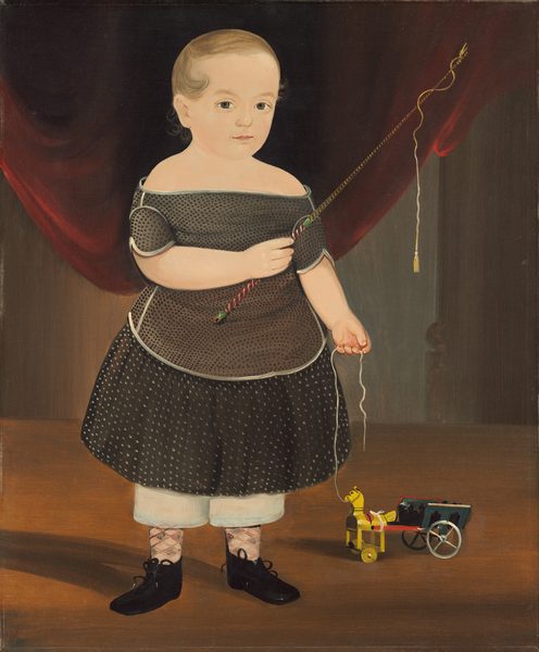 The image shows a young child standing upright. The child has light-colored, short hair, a pale complexion, and delicate features. They are dressed in a dark garment with a dotted pattern and light edges, light-colored pantaloons, patterned socks, and dark shoes with bows. In their right hand, the child holds a stick with a string attached, and in their left hand, they hold a string connected to a small colorful toy horse pulling a cart. The backdrop consists of red curtains on the sides, against a wooden floor and wall.