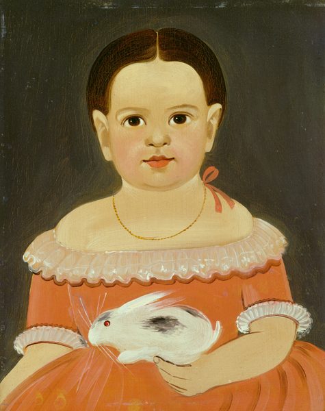 The image is a portrait of a young child shown from the waist up. The child is seated and facing forward. The child has dark hair that is styled smoothly and parted down the middle. The facial features include wide, dark eyes and a small, full-lipped mouth. The child is wearing a dress with a wide neckline and puffed sleeves, which is coral in color with delicate white lace trim. Around the child's neck is a simple gold chain necklace. The child holds a white rabbit with a black spot on its back in their right arm. The background is a plain, dark gray color.
