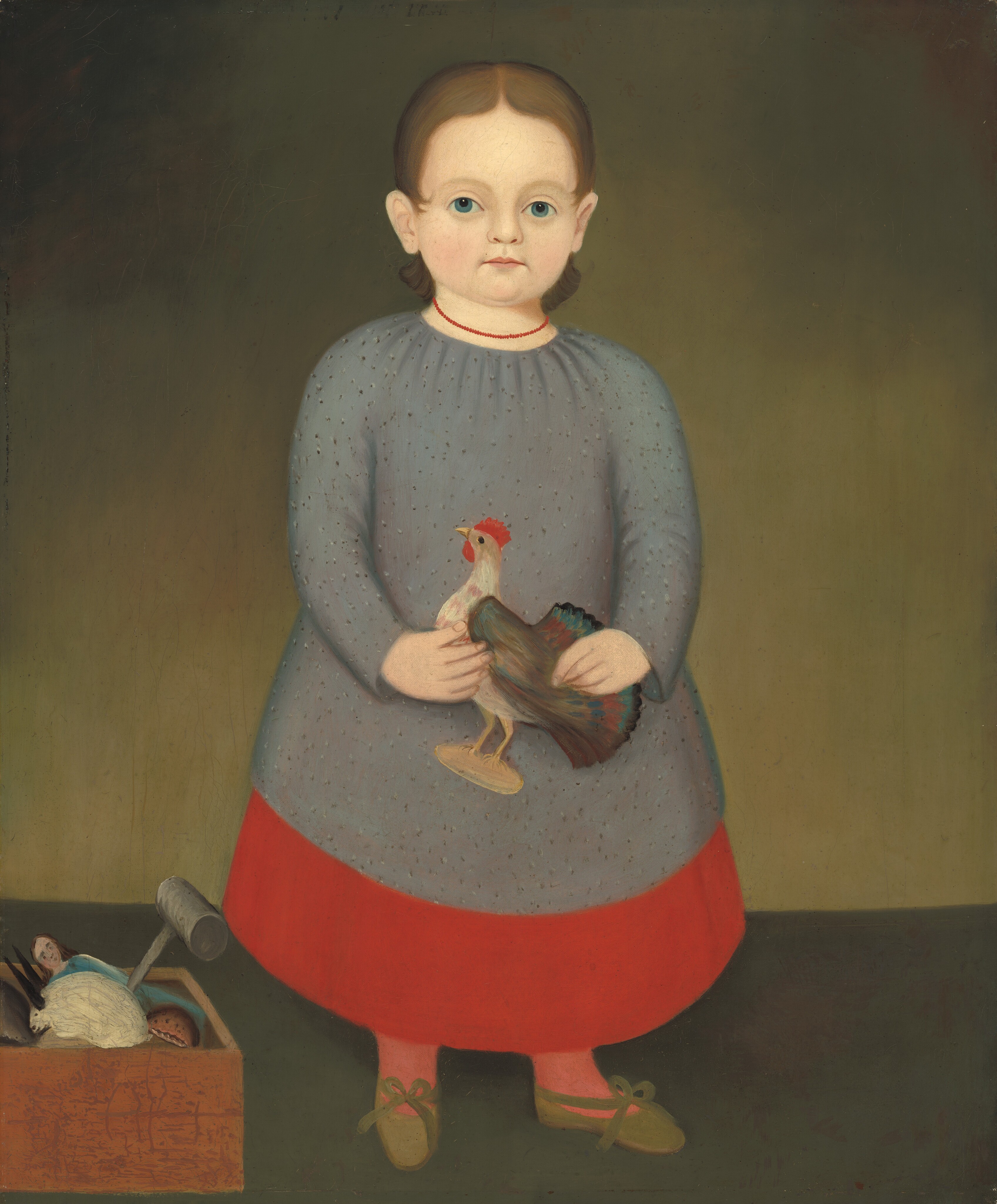 The image shows a young child standing upright, facing forward. The child has light skin, light brown hair, light blue eyes, and a neutral expression. They are dressed in a long, gray dress with a red lower part, red socks, and brown shoes with green bows. The child is holding a small model of a rooster. Next to them, on the floor, is a box with small objects like a doll and a ball. The background is a simple greenish-brown gradient.
