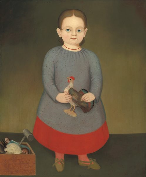 The image shows a young child standing upright, facing forward. The child has light skin, light brown hair, light blue eyes, and a neutral expression. They are dressed in a long, gray dress with a red lower part, red socks, and brown shoes with green bows. The child is holding a small model of a rooster. Next to them, on the floor, is a box with small objects like a doll and a ball. The background is a simple greenish-brown gradient.