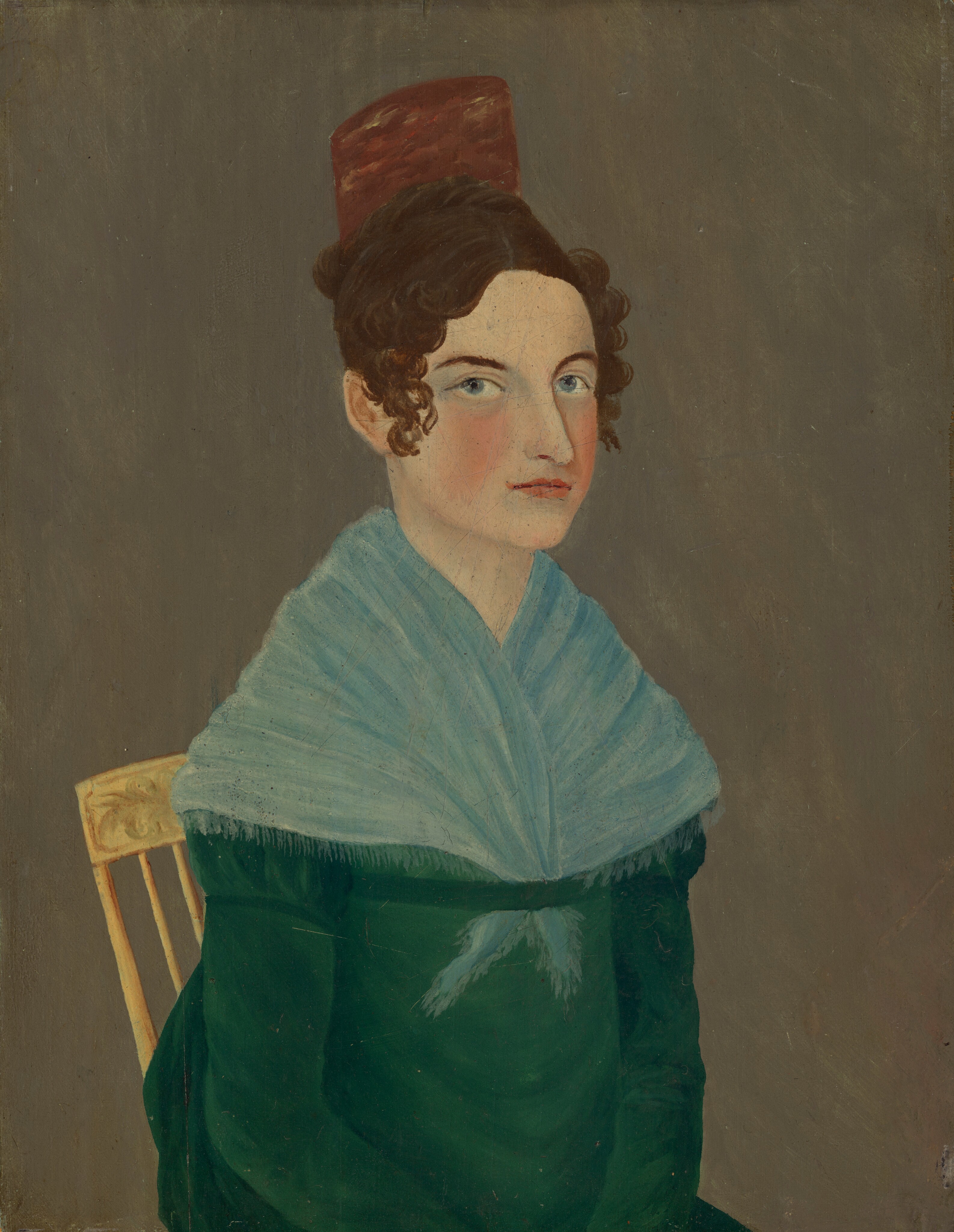 The image depicts a person sitting on a wooden chair with a decorative backrest. The individual is facing slightly to the right, making eye contact with the viewer. They have fair skin, blue eyes, reddish lips, and dark curly hair styled with a wave, partially covered by a brown fez-like hat. The person is wearing a dark green garment underneath a light blue fringed shawl. The background is a plain, muted brown color.