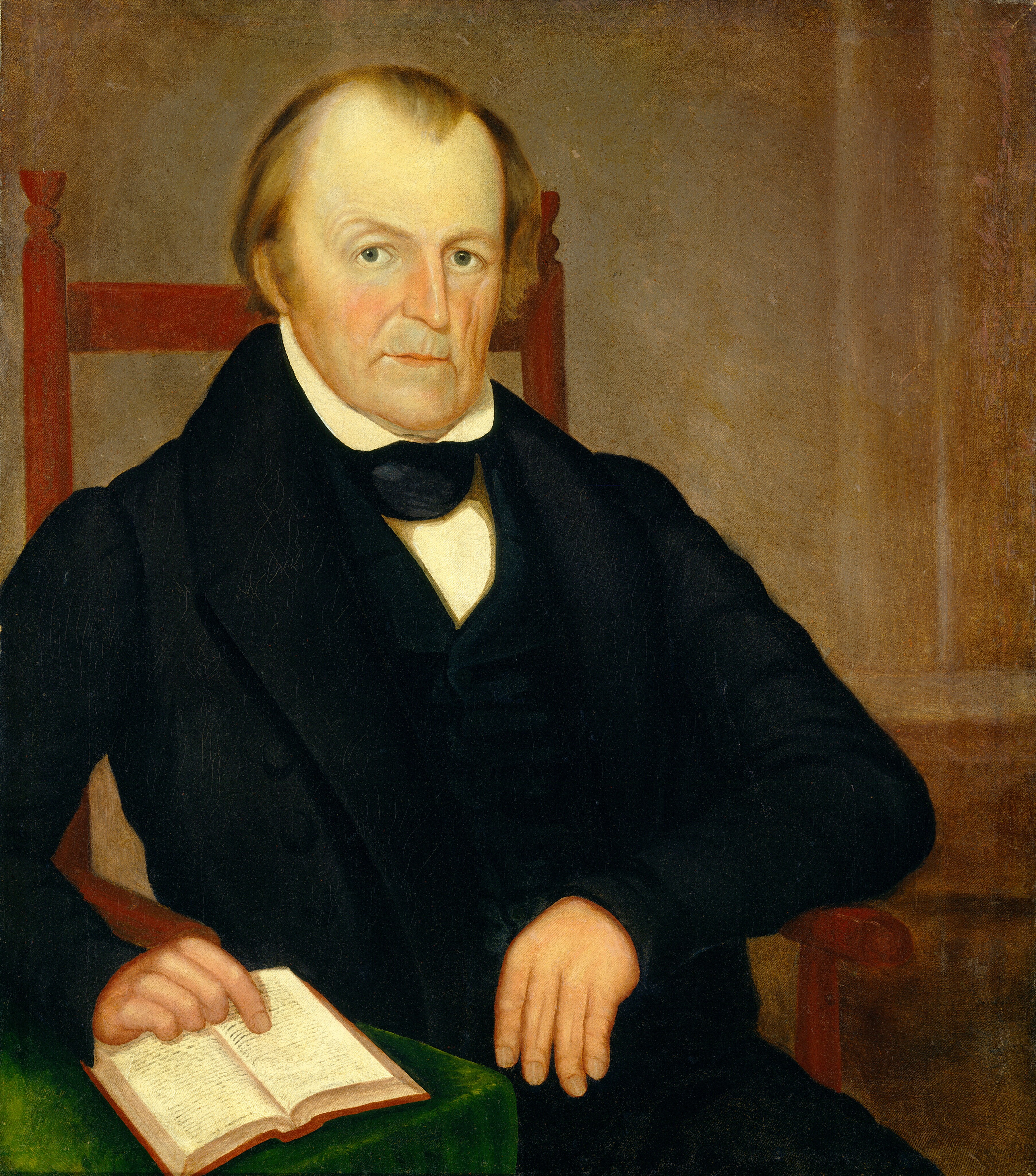 The image depicts a man sitting in a chair. He has fair skin, light brown hair, and is wearing formal attire. He is gazing directly at the viewer. The man is holding an open book in his left hand, while his right arm rests on the chair's arm. The background is plain and brown, drawing attention to the man.