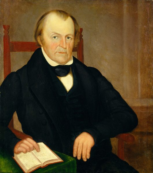 The image depicts a man sitting in a chair. He has fair skin, light brown hair, and is wearing formal attire. He is gazing directly at the viewer. The man is holding an open book in his left hand, while his right arm rests on the chair's arm. The background is plain and brown, drawing attention to the man.