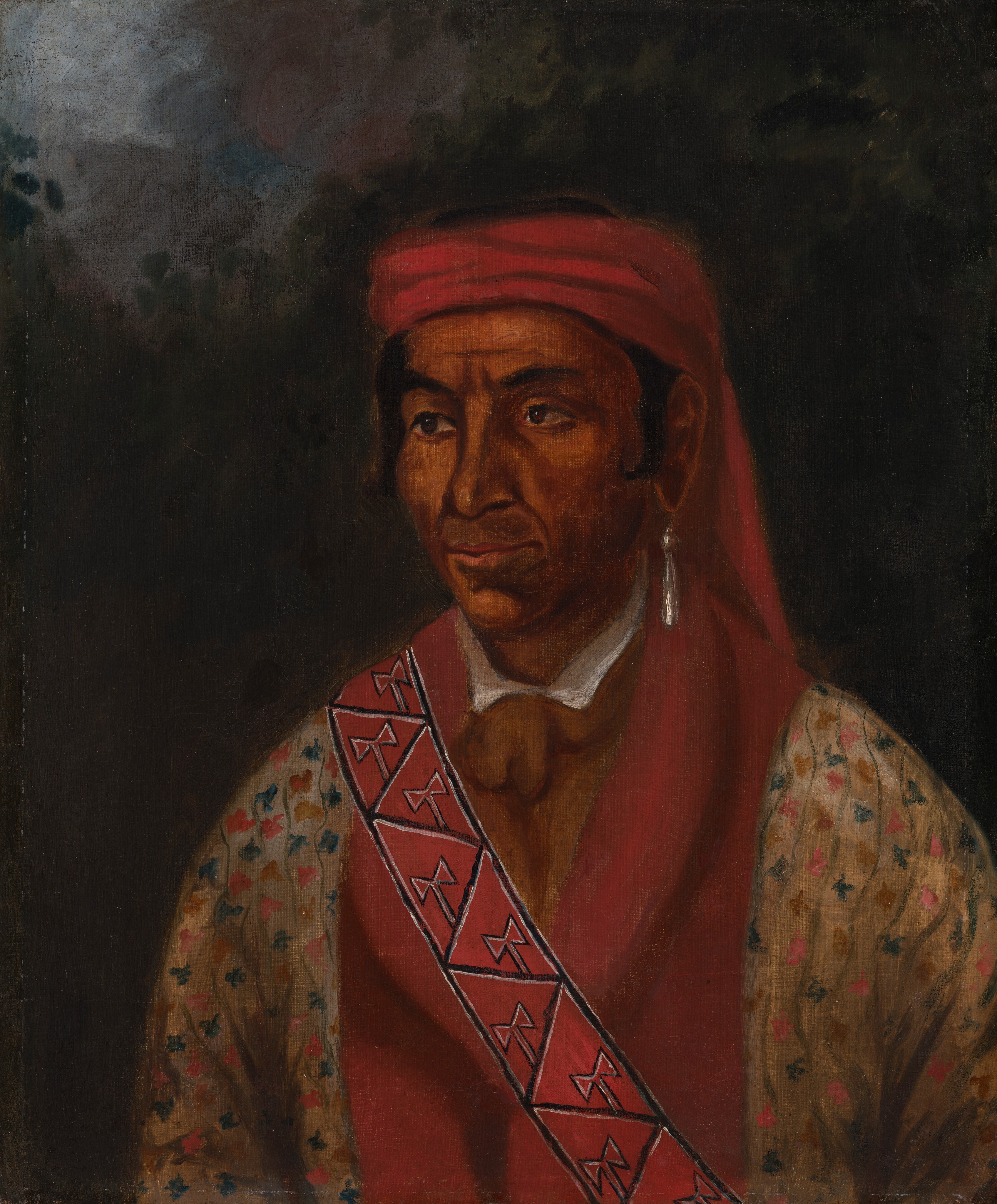 The image shows a man's upper body from the chest up. He is positioned slightly to the side, looking to the left. The man has a strong jawline, dark hair mostly covered by a red headwrap, deep brown skin, a shirt with colored flower patterns, a red sash with geometric designs, and a long silver earring on his left ear. The background is dark with no discernible objects.