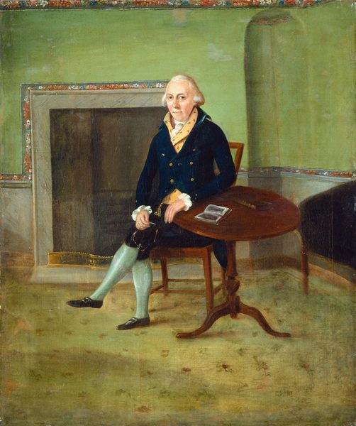 The image is a full-body portrait of a seated man. He is positioned slightly turned to his left with his legs crossed. His facial features show a calm expression, and he has light skin and white hair that is styled backward. The man is wearing formal attire that includes a dark blue coat with large gold buttons, a yellow waistcoat, a white neck scarf with floral patterns, knee-length black breeches, and light blue stockings. He sits on a wooden chair next to a round wooden table, on which there is an open book and a small rectangular object. The background features a green wall with an ornamental border and a large, empty fireplace behind him. The floor is covered with a light-colored carpet that has a simple pattern.