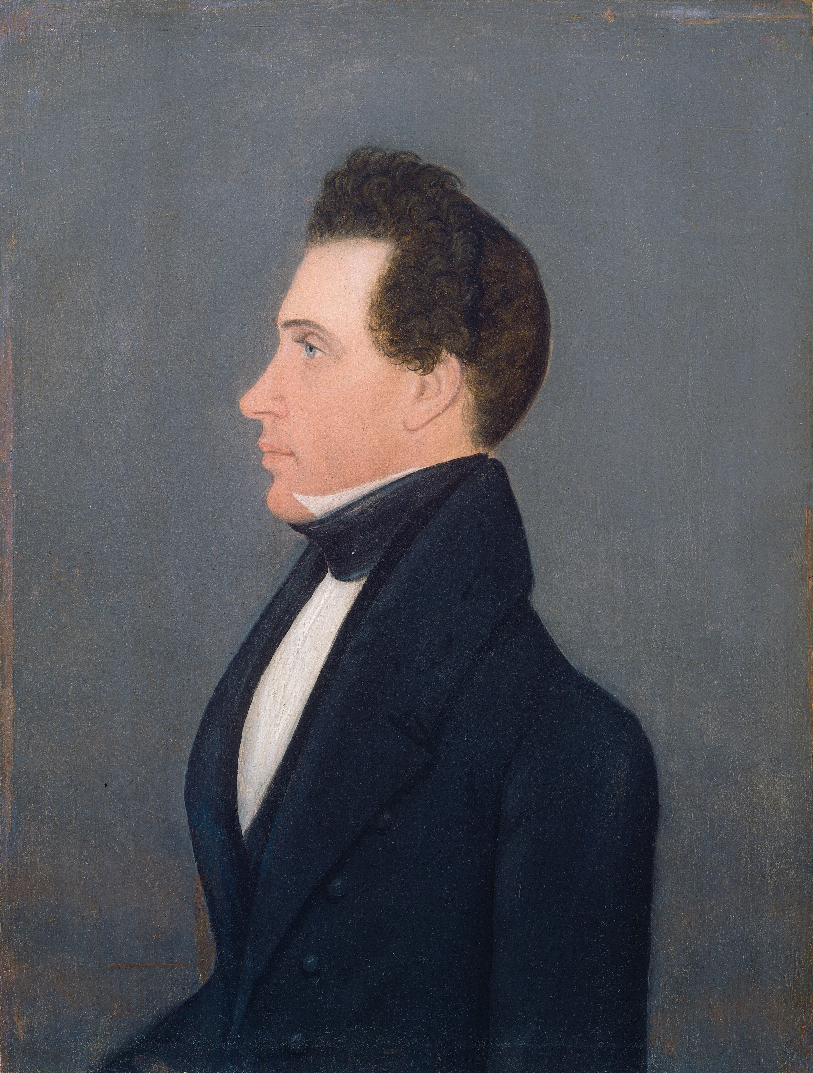 The image shows the upper half of a man's body in profile view facing left. The man has a straight posture, with prominent facial features including a nose and chin. His hair is dark, curly, and close to his head, with some volume on top. He wears a dark suit, high-collared white shirt, and black cravat. The background is plain grayish-blue with no visible objects.