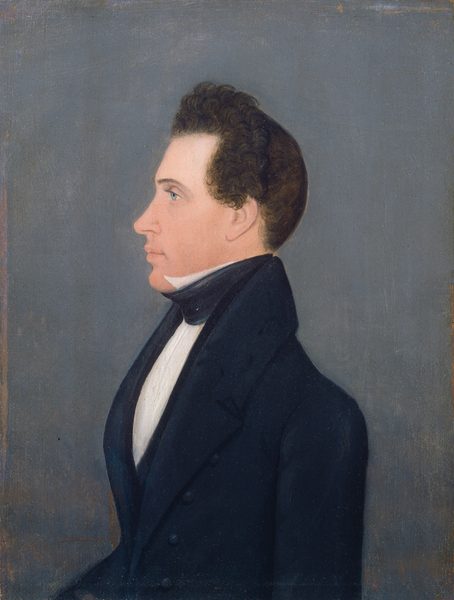 The image shows the upper half of a man's body in profile view facing left. The man has a straight posture, with prominent facial features including a nose and chin. His hair is dark, curly, and close to his head, with some volume on top. He wears a dark suit, high-collared white shirt, and black cravat. The background is plain grayish-blue with no visible objects.
