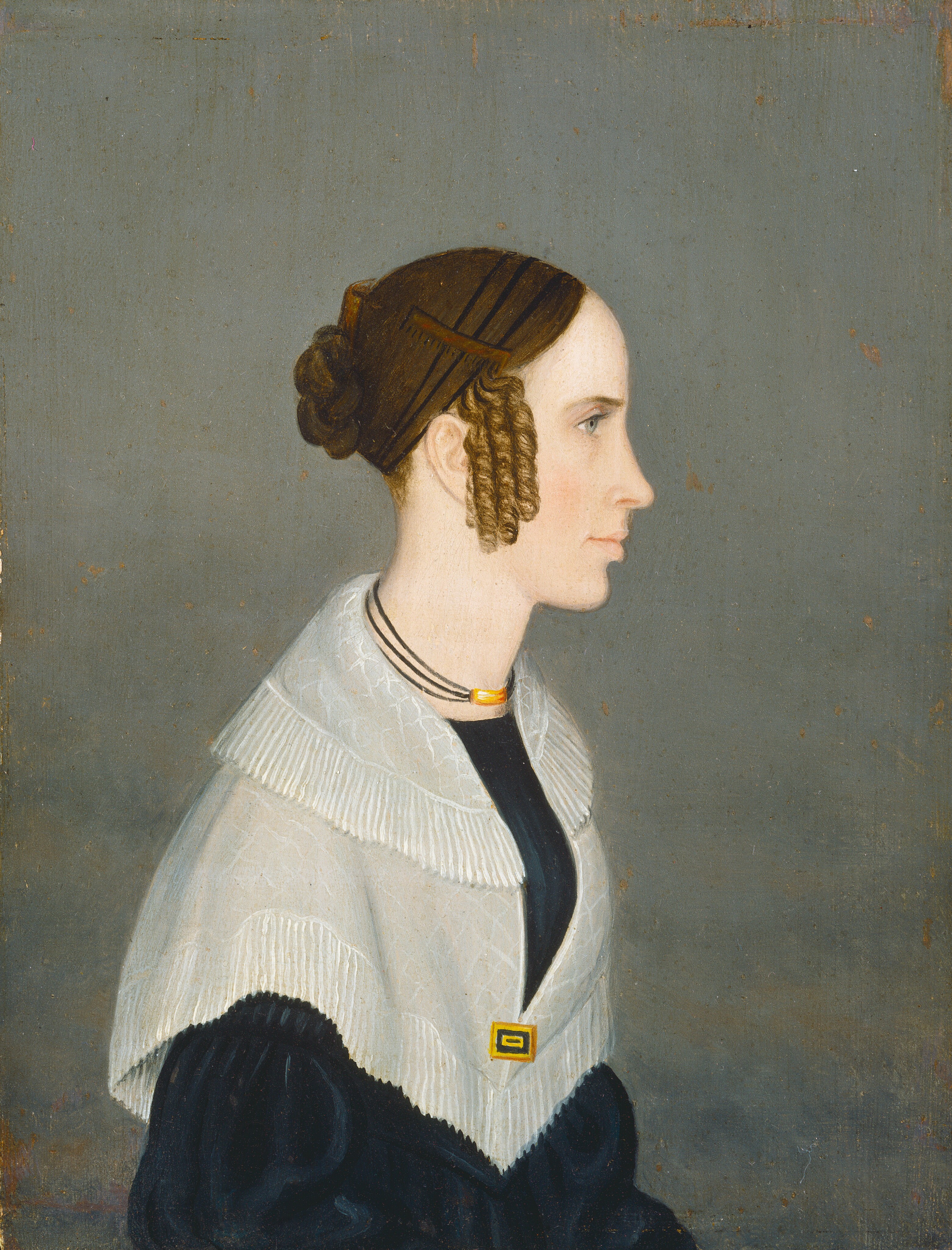 The image shows the upper half of a person in profile, facing to the right with a slightly turned head. The person has fair skin, straight nose, and a slight smile. The brown hair is styled in an updo with tight curls around the face. They wear a black garment with a light shawl, a rectangular clasp, and a triple-strand necklace with a central gold or amber pendant. The background is a simple, muted gray.