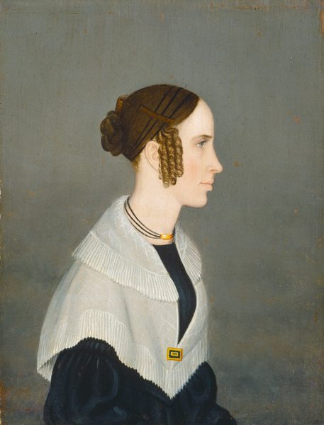 The image shows the upper half of a person in profile, facing to the right with a slightly turned head. The person has fair skin, straight nose, and a slight smile. The brown hair is styled in an updo with tight curls around the face. They wear a black garment with a light shawl, a rectangular clasp, and a triple-strand necklace with a central gold or amber pendant. The background is a simple, muted gray.