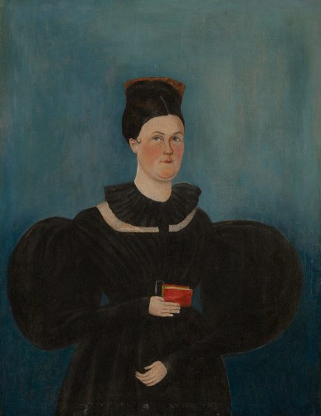 The image shows a woman standing upright with a small red book in her right hand. She has a fair complexion, neutral expression, slightly arched eyebrows, and small, rounded lips. Her dark brown hair is styled in an updo with a black accessory. She wears a high-necked dark dress with puffed sleeves and a white collar. The background is plain blue. The woman is not wearing any visible jewelry, and the setting is minimalistic with only the book present.