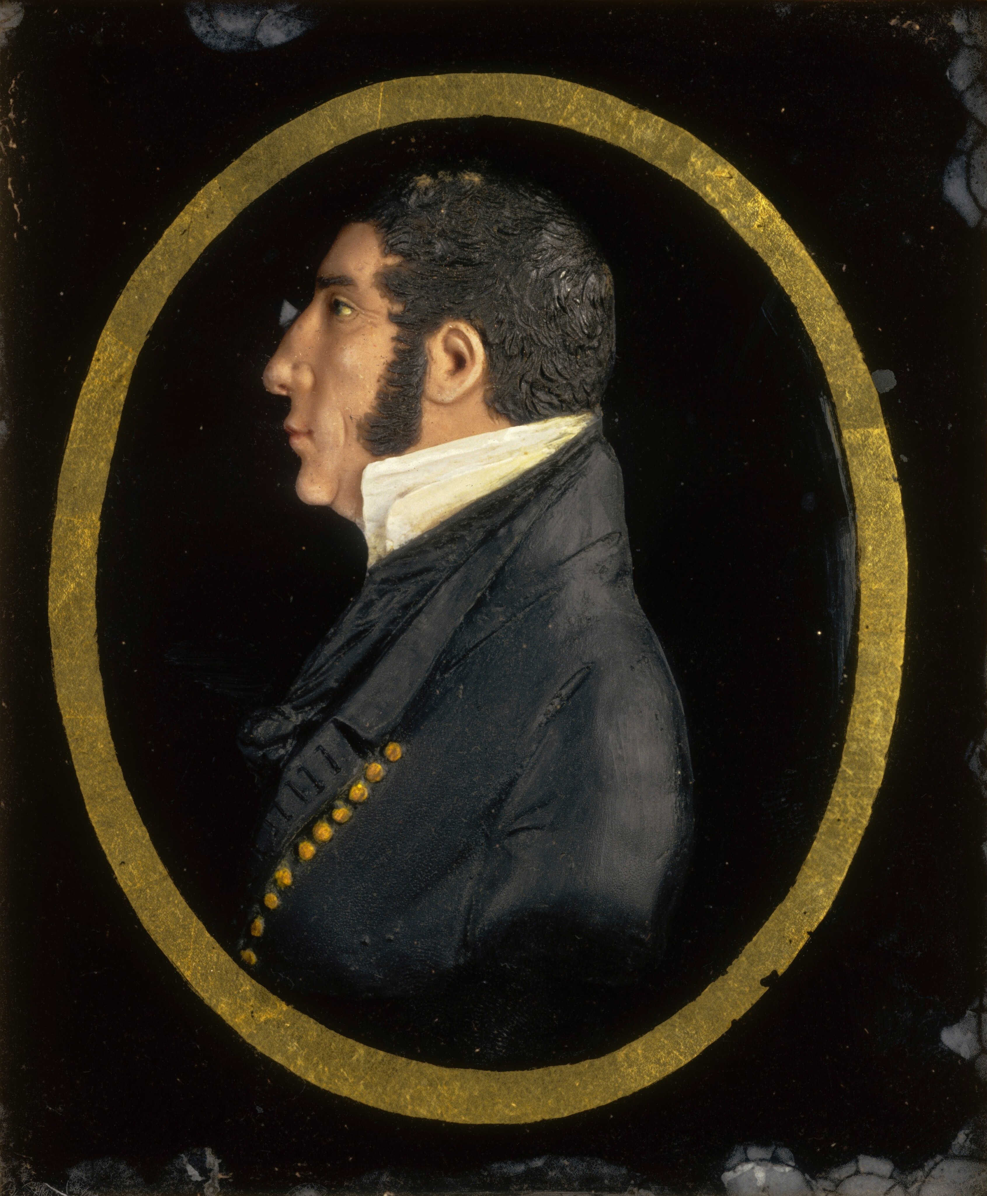 The image depicts a portrait miniature of a man's upper body in profile facing left. The man has defined facial features, a prominent nose, sideburns, dark hair in waves, and wears formal attire with a high, white collar and gold buttons. The background is solid black with a gold oval border around the man.