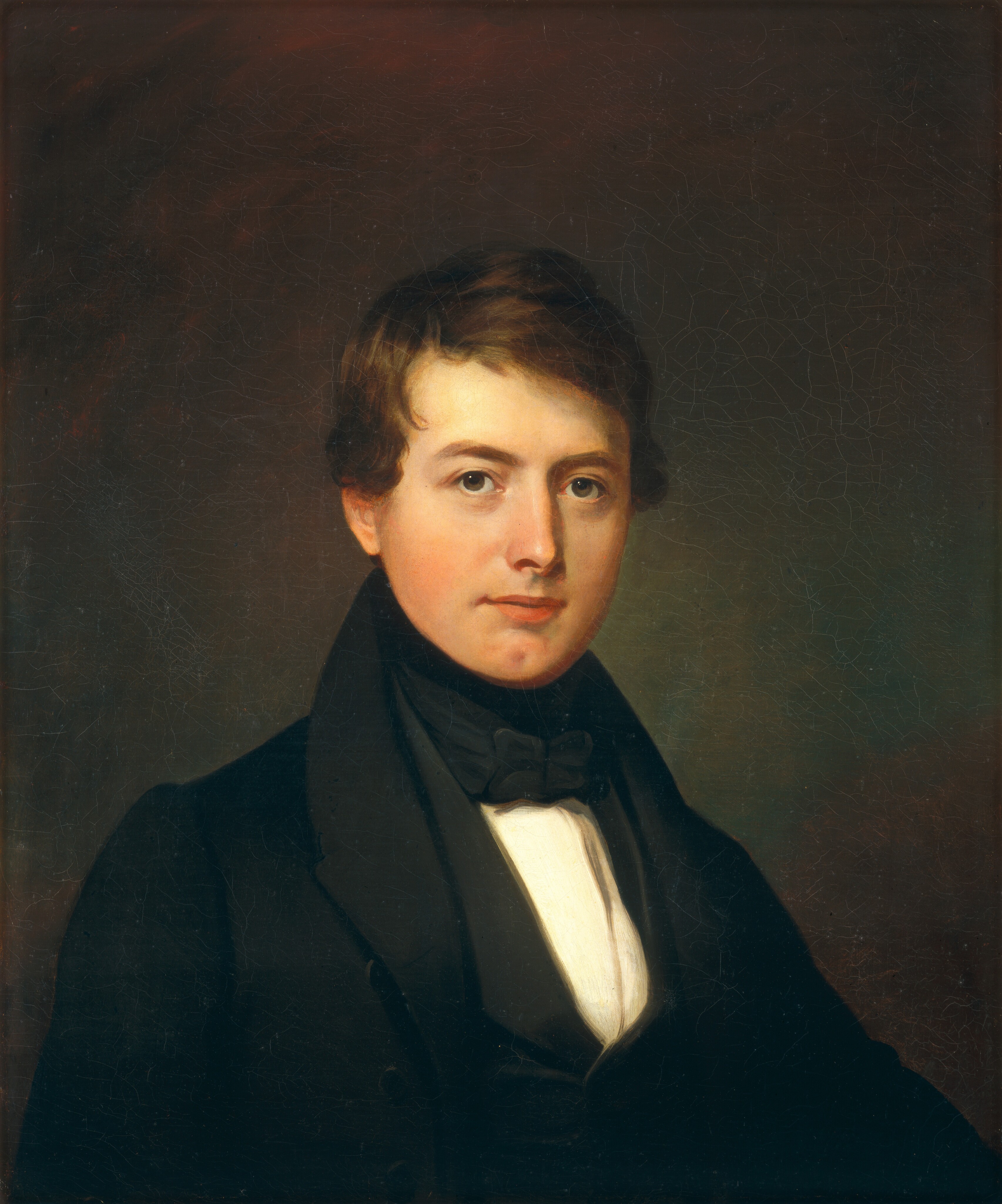 The image depicts a man's upper body in a classic portrait pose. He faces forward, looking at the viewer. The man has facial features like fair complexion, a smooth face, warm brown eyes, and styled hair with a slight wave and a side part. He wears formal attire with a dark jacket, white shirt, black waistcoat, and a black bow tie. The background is a plain, muted color with no visible jewelry or objects.