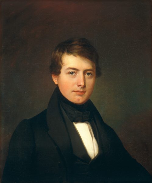 The image depicts a man's upper body in a classic portrait pose. He faces forward, looking at the viewer. The man has facial features like fair complexion, a smooth face, warm brown eyes, and styled hair with a slight wave and a side part. He wears formal attire with a dark jacket, white shirt, black waistcoat, and a black bow tie. The background is a plain, muted color with no visible jewelry or objects.
