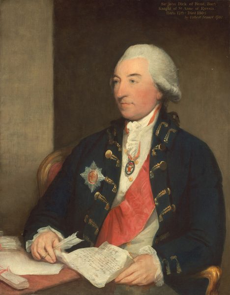 The image depicts a man sitting, facing slightly to the right. He has fair skin, a straight nose, and white, slightly curly hair styled in an 18th-century manner. He wears a navy blue coat with gold embellishments, a white shirt with a ruffled collar, a red sash, and a star-shaped badge. A medallion hangs around his neck. The man is seated at a table with a document in front of him, holding a quill pen. The background is neutral.