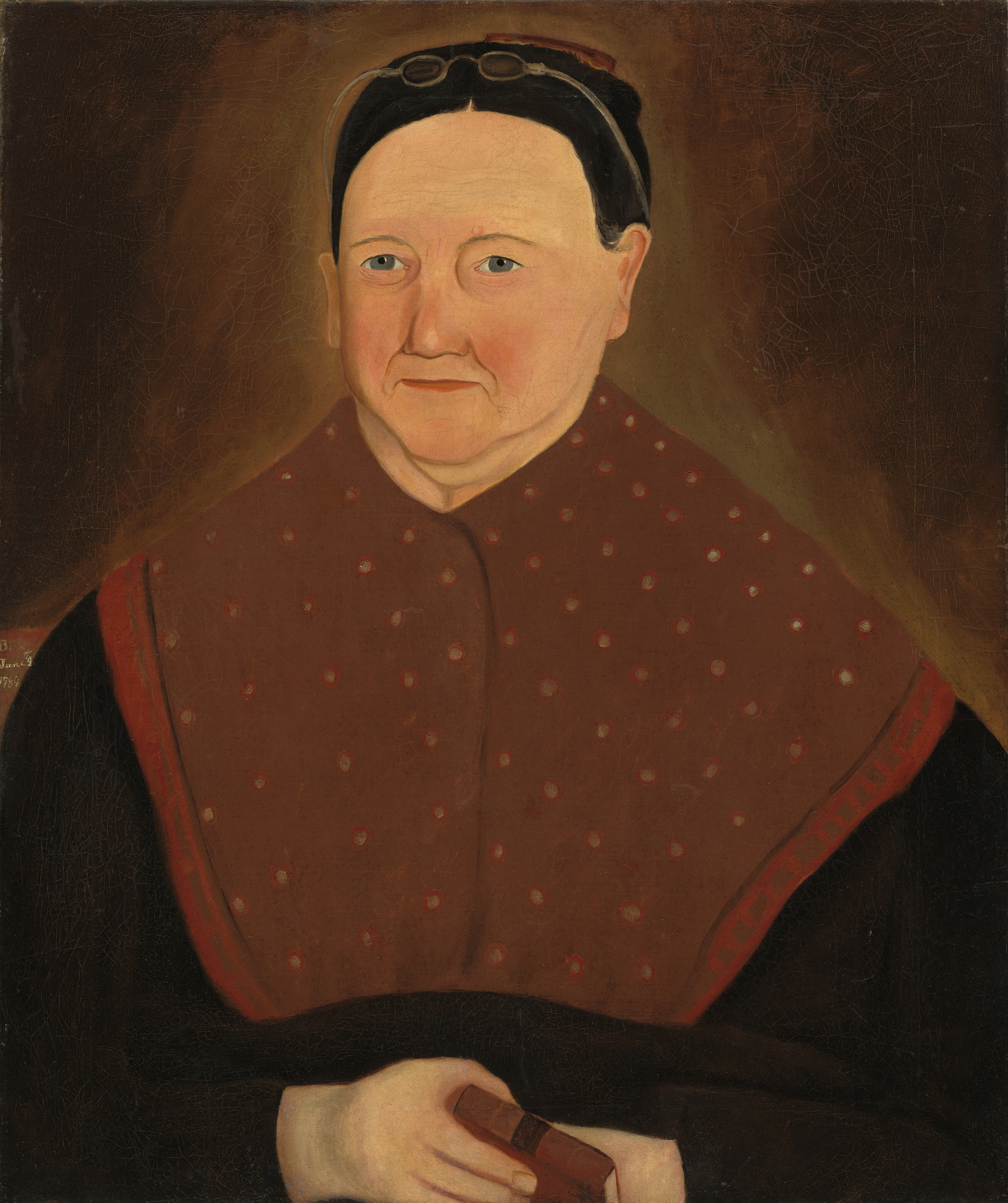 The image depicts an elderly woman seated, holding a closed book with her hands folded. She has light skin, small blue eyes, and a slight smile. Her hair is dark and smooth, with round spectacles on top of her head. She is wearing a dark garment with a brown shawl featuring red dots. The background is muted brown, emphasizing the woman.
