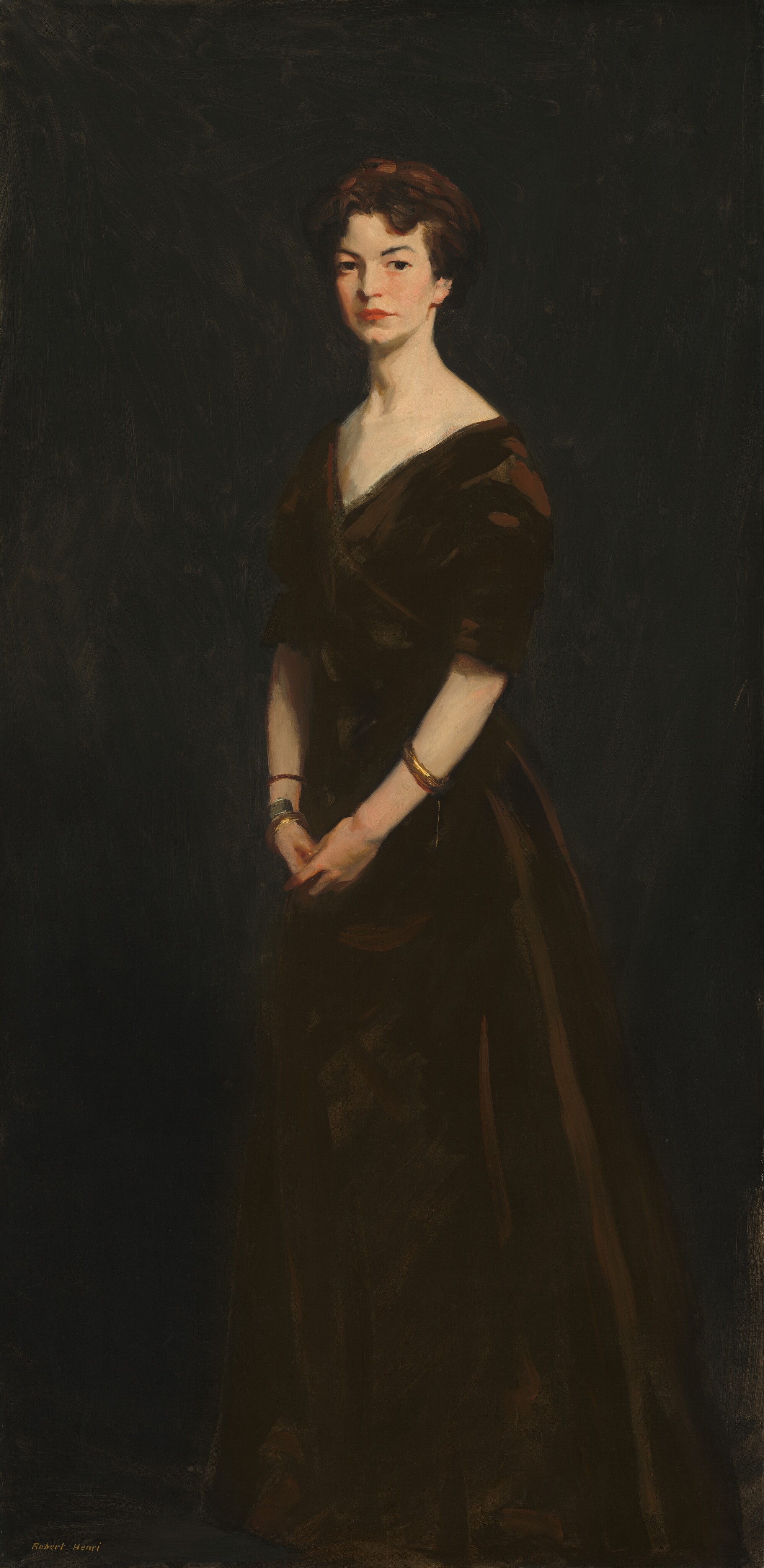 This painting is a full-body portrait of a woman. She is standing in an upright position with her hands clasped together in front of her body. Her body is faced slightly to the left, but her face is turned to us. Her face is serious, with pale skin, dark, arched eyebrows, a cleft chin, and red lips in a neutral expression. She has short, dark, softly curled hair. She wears a dark dress with wide shoulders, a V-shaped neckline, and elbow-length sleeves, accessorized with gold bangles on her wrists. The background is a dark, almost black, monochromatic backdrop that blends in with the bottom of her skirt.