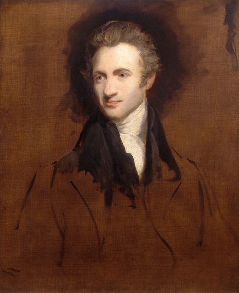 The image shows a man with light complexion in a formal outfit against a plain background. He is turned slightly to his left, with well-defined facial features including prominent cheekbones, a straight nose, and slightly upturned lips. His hair is voluminous, brushed back and slightly wavy. He is wearing a high-collared white shirt with a black jacket.