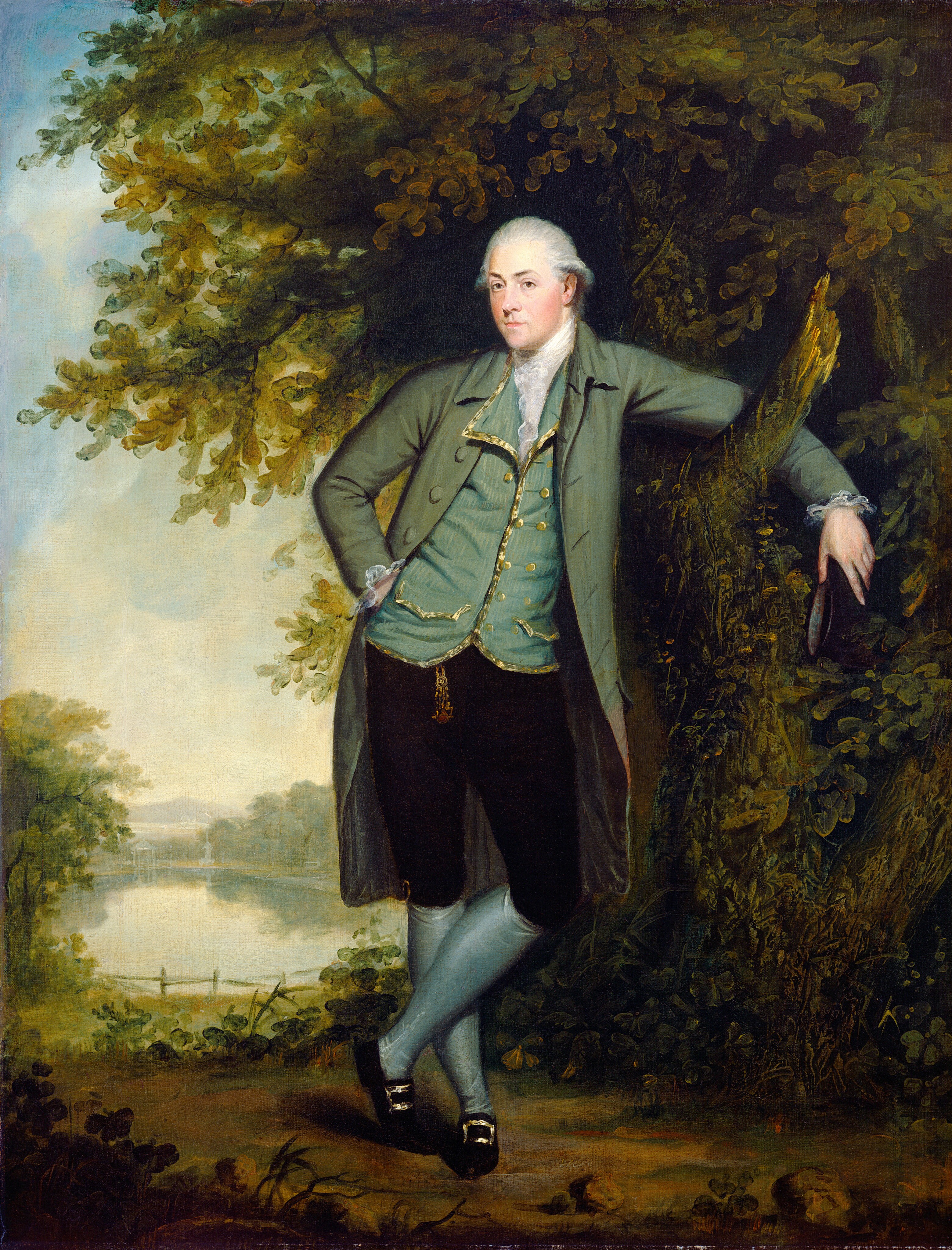 The image displays a man in 18th-century attire standing against a tree. He has fair skin and powdered white hair. He is elegantly dressed in a green coat, blue waistcoat, white cravat, dark breeches, blue stockings, and black shoes with gold buckles. The man is holding a three-cornered hat, standing with one arm on a tree branch, legs crossed at the ankle, with a calm expression and a slight smile. The background features a tree, serene landscape with foliage, a reflective body of water, distant hills, and a cloudy sky.