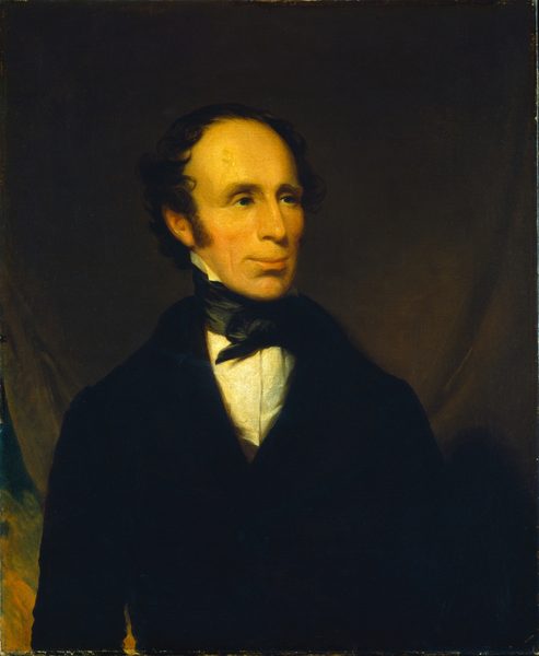 The image is a portrait of a man's upper body. The man is seated, facing to his left with a direct gaze. He has fair skin, a slender face with a prominent forehead, long nose, and slight smile. His dark, curly hair is receding at the temples. He wears a black coat, white shirt, and black necktie. There are no visible accessories. The background is a dark, muted brown color.