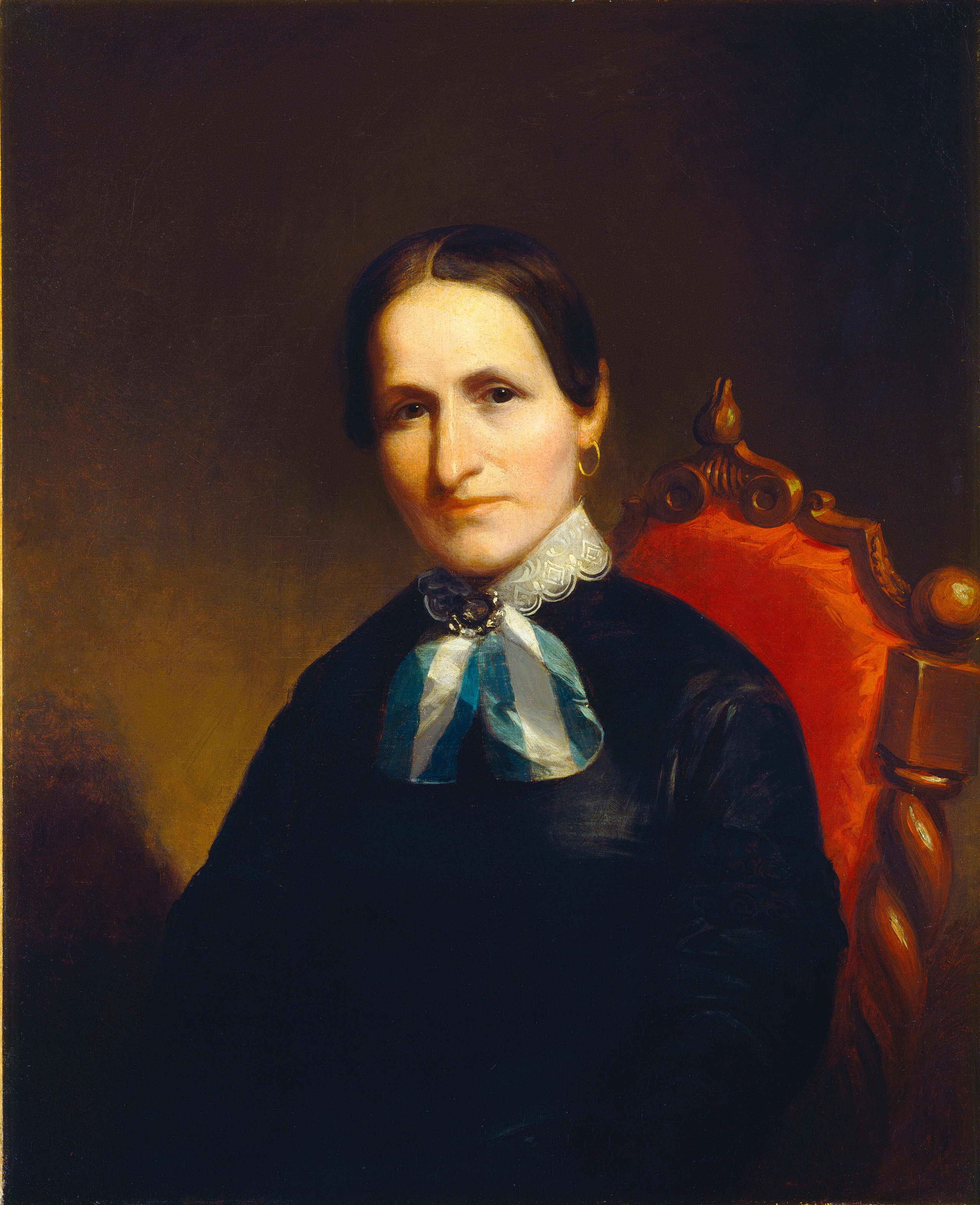 The image shows a portrait of a woman from the waist up, seated in a three-quarter view facing slightly to her right. Her facial features include high cheekbones, a straight nose, and brown eyes. Her hair is parted in the middle and smoothed back tightly, revealing her ears. She is wearing a dark dress with a high collar and white lace trim. Around her neck is a large bow or tie with light and dark blue stripes, accented by a brooch at the center. She has gold hoop earrings. The chair she sits in has a rich red upholstery with ornate wooden carvings. The background is a muted dark color.