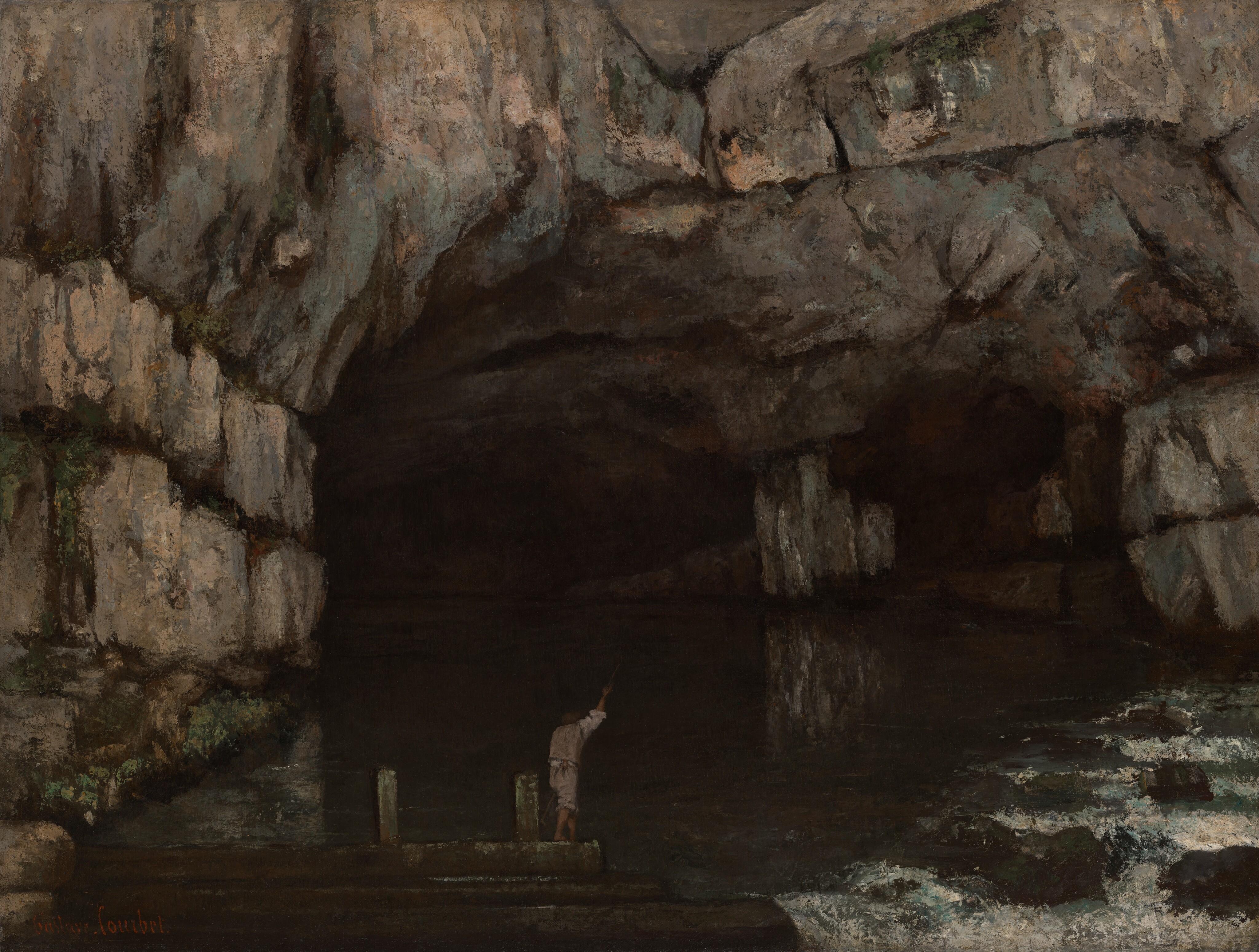 A pair of arched cave openings yawn in a steep, rocky cliff face that fills this horizontal painting. The cliff face is painted with dabs and textured strokes of slate gray, peachy tan, and muted blue, and some moss grows along crevices to our left. The opening to our left of center is four times the size of the smaller opening next to it. Murky green water reflects the shadowy openings and the pier-like stone between them. The water churns white over large boulders to our right, in the lower corner of the canvas. A pale-skinned man, minuscule in scale in front of the larger opening, stands barefoot with his back to us. His pants are rolled up over his knees and the long shirt sleeves up to the elbow under a vest. One hand is raised to hold a long stick. He is on what might be a pier stretching across the water. The artist signed the work with red paint in the lower left corner, “Gustave Courbet.”