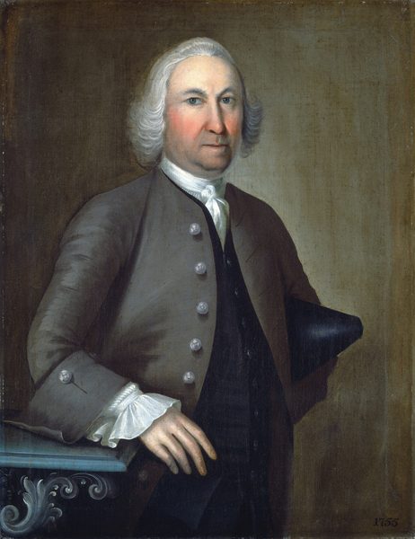 The image shows an upper-body portrait of a man standing in a slight profile facing the viewer's left. He has a serious expression with a clean-shaven face, strong facial features, and white hair styled to the sides. He wears a dark coat with large buttons over a white shirt featuring ruffled cuffs and a dark waistcoat underneath. His left hand rests on an ornate piece of furniture, which displays intricate carvings. The background is a plain, textured wall. The date "1755" is inscribed in the lower right corner of the painting.