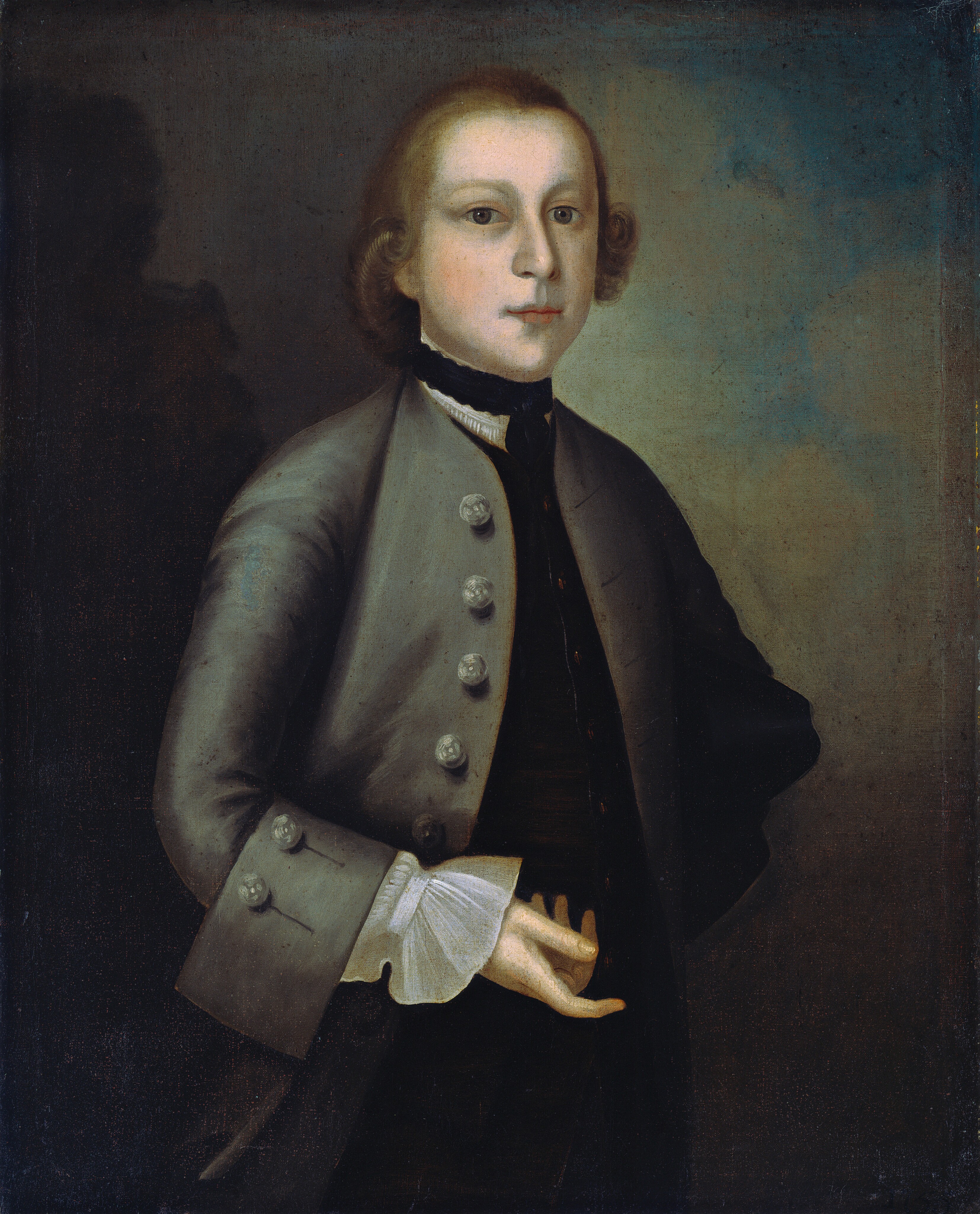The image shows a young man with fair skin and a rounded face, wearing an 18th-century style outfit. He has light brown curly hair, a gray coat with large buttons, black collar, and white neckcloth. He stands with his right hand in his coat and his left hand slightly visible. The background is a combination of dark gray and light brown tones.