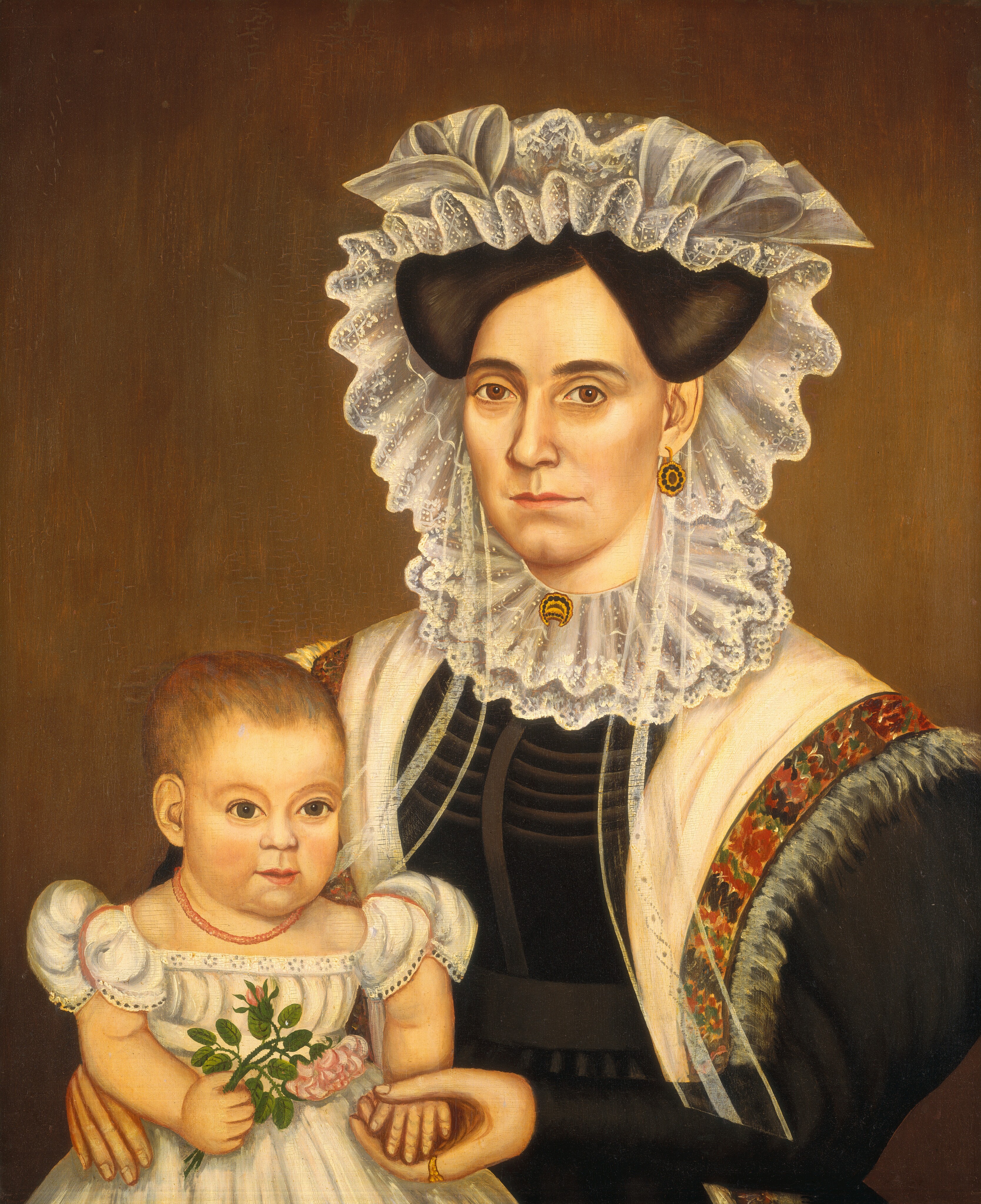 The image shows an upper body portrait of a woman and a child. The woman is seated and faces the viewer, with a serious expression. She wears a dark dress with a high neck and elaborate detailing, a shawl with ornate embroidery over her shoulders, gold earrings, and a brooch. Her hair is styled under a white laced cap. The child, with light hair, sits on the woman's lap, facing the viewer, wearing a white dress with lace details and a red bead necklace. The child holds a bouquet of flowers in one hand. The background is plain and dark.
