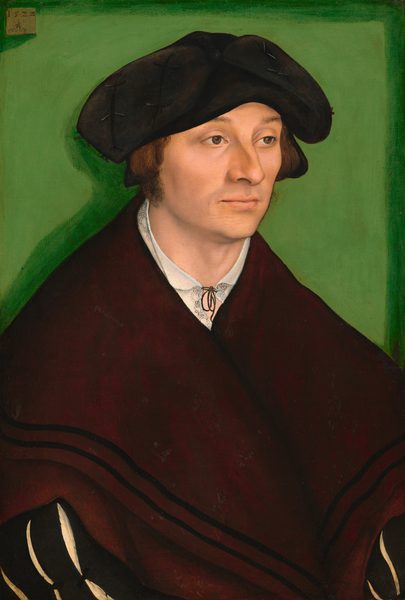Shown from the waist up, a clean-shaven man with pale, peachy skin is dressed in black and burgundy red in this vertical portrait painting. His shoulders are as wide as the composition and his hat nearly touches the top edge, so he almost fills the painting. His body and face are angled to our right, and he looks off in that direction with hooded brown eyes under slightly raised brows. He has a long, bumped nose, high cheekbones, the hint of a five-o-clock shadow, and his pale pink lips are closed. His hair is cut straight across at ear length, and fuzzy sideburns come down to his jawline. A wide, soft black cap curves down either side of his head to ear level. He wears a white shirt with a delicate black pattern on the neckline, which is tied with a thin black ribbon. A voluminous dark red shawl or wrap has two black bands along the bottom hem, and it engulfs his sloping shoulders. In the lower corners of the painting, we see long black sleeves under the wrap. The sleeves have slashes to allow the white undergarment to show through. He is set against a lime-green background and illuminated by bright light from the upper right, which creates a darker green shadow behind him, to our left. The artist signed the painting with a symbol on a small, light brown panel, which appears to be affixed to the back wall in the upper left corner of the painting. The date, “1522,” appears over a winged serpent holding a ring in its mouth.