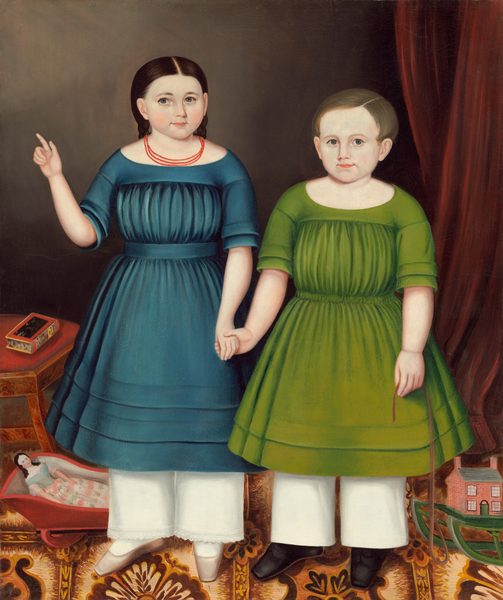 The image features two young children standing side by side. The child on the left extends one arm sideways and has the other arm lowered, while the child on the right has both arms relaxed by their sides, one holding a string. Both children have round faces with light complexions; the child on the left has dark brown hair styled in waves down to the shoulders, while the child on the right has light brown hair. The child on the left is dressed in a blue dress with a loosely gathered waist and short sleeves, white stockings, white shoes, and a red beaded necklace. The child on the right wears a green dress with similar design features, white stockings, and black shoes. Near their feet are a toy doll in a red cradle and a small toy house. In the background, there is a plain dark wall, a deep red drapery, a carpet with elaborate patterns, and a small wooden table with a box on top.