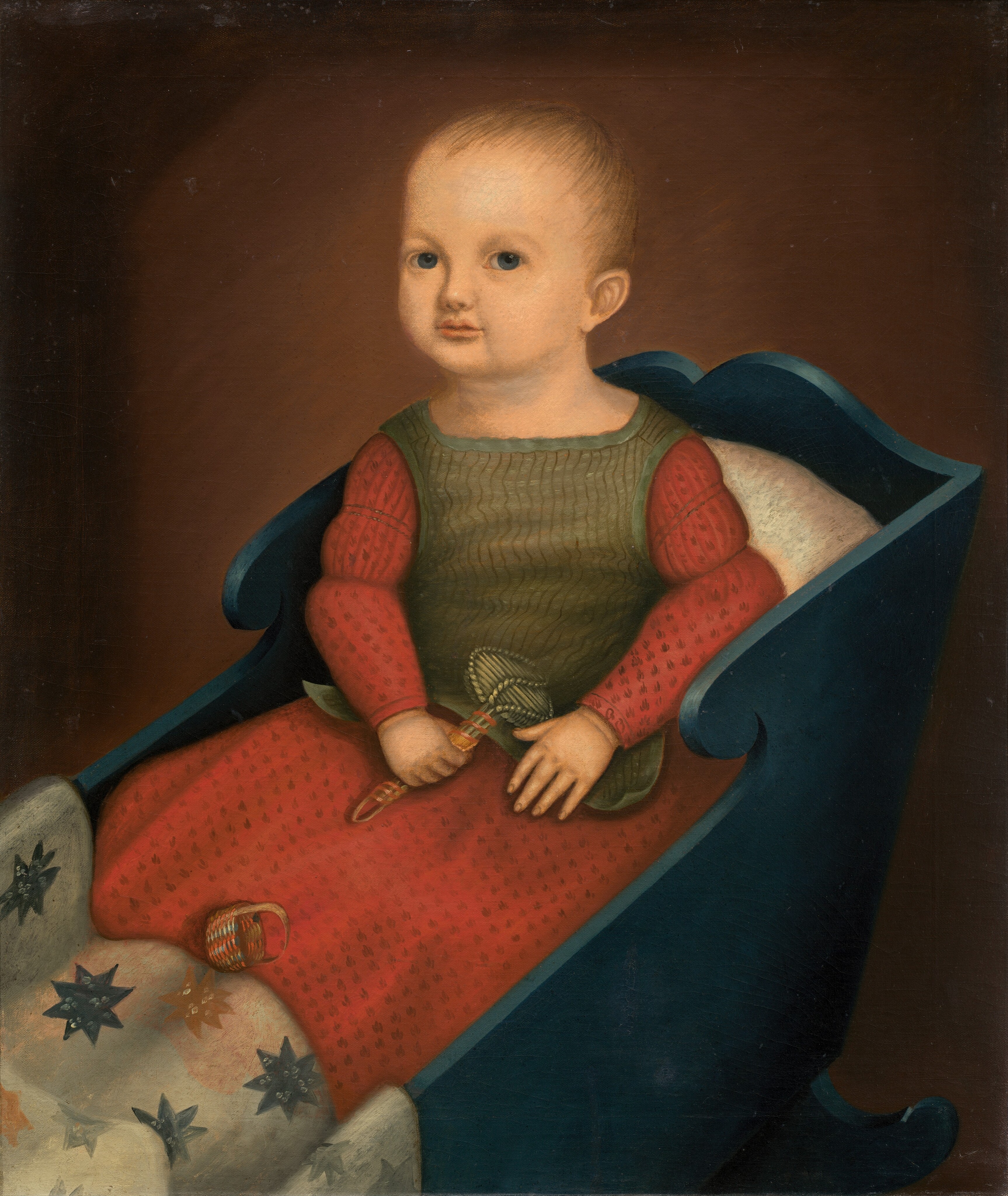 The image depicts an infant seated in a cradle. The child is sitting upright and facing slightly to the side. The infant has short, light-colored hair and wears a red long-sleeve dress with black patterns, covered by an olive-green vest. The cradle is made of dark blue wood and there is a cream-colored cushion inside. The child holds a rattle and there is a small woven basket beside them. The background is plain brown.