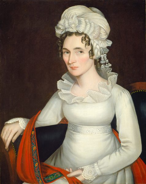 The image depicts a woman seated from the waist up. She faces forward with her body slightly angled to her left. Her facial features include arched eyebrows, bright eyes, and a neutral expression. Her hair is styled with soft curls framing her face and is mostly hidden by a white headpiece with lace and fabric detailing. She wears a white dress with lacework on the neckline and sleeves, along with a white belt. A red shawl with a colorful border is draped over the chair, held by her right hand. The background is a solid dark color, emphasizing her as the main subject.