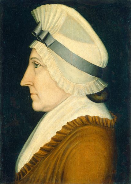 The image shows the profile of a person from the chest up, positioned in a side view orientation facing left. The person has a prominent straight nose, deep-set eyes, and distinct eyebrows. Their hair is covered by a large white cap with pleats and a blue-gray ribbon. They are wearing a brown garment with a high, white pleated collar. The background is a dark, solid color.