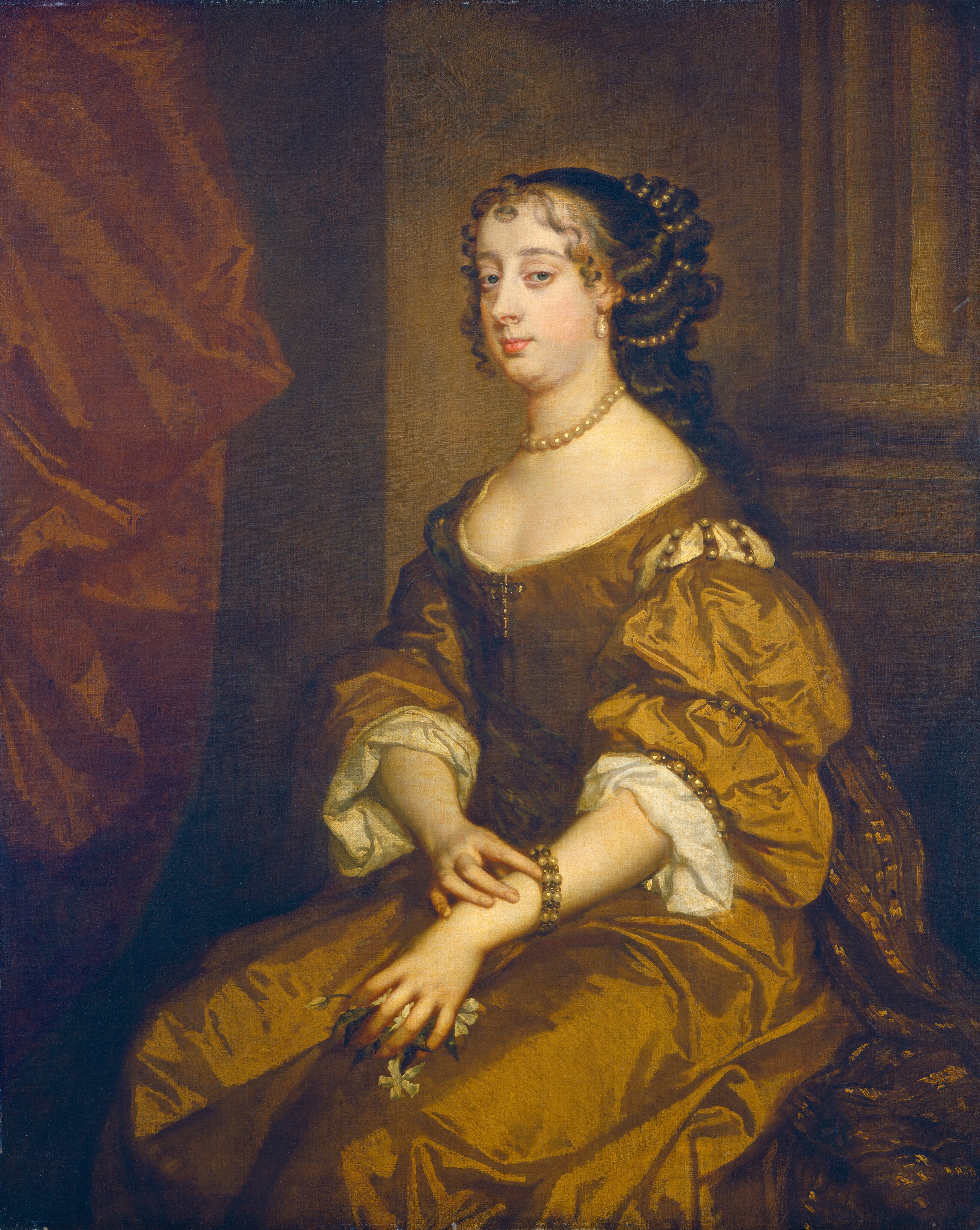 This painting is a portrait of a woman. She is seated and facing slightly to the left, with her hands in her lap. She has fair skin, blue eyes, a soft chin, dark curly hair, and pink lips in a neutral expression. She is wearing a golden-brown gown with voluminous sleeves, a low neckline, and pearl accents. She is also adorned with a pearl necklace, earrings, and a bracelet, with more pearls in her long hair. In one hand she is holding white flowers. The background is a warm, dark-toned interior with some red drapery visible on the left.