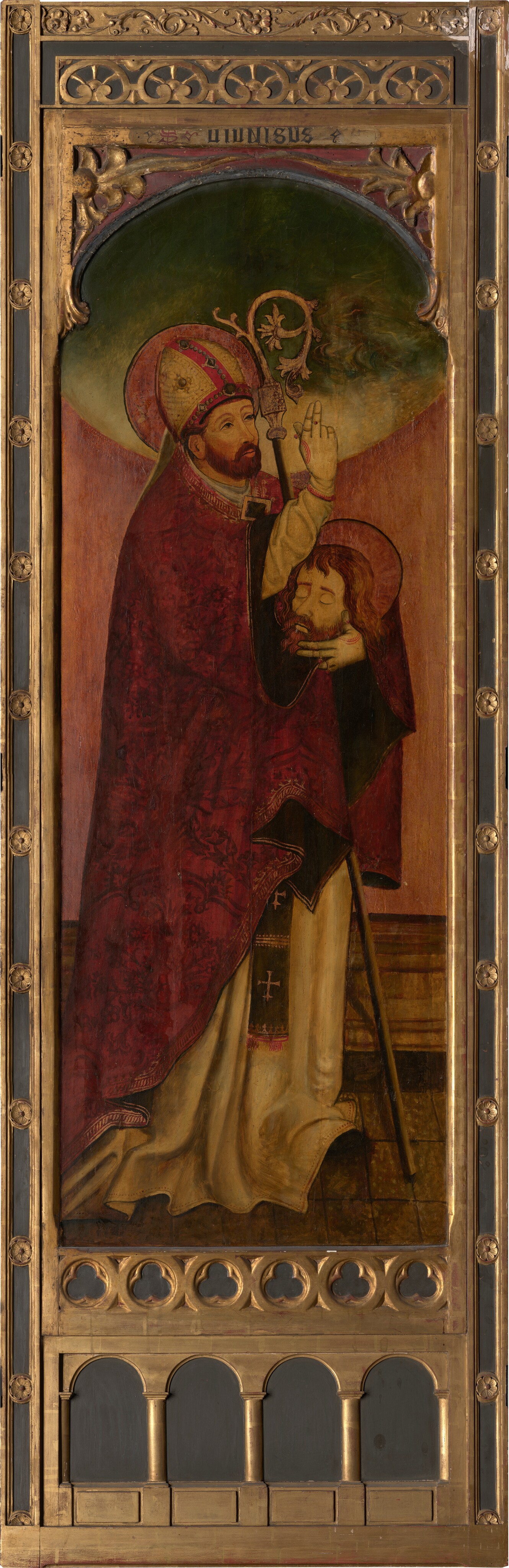 The image shows the upper half of a standing figure. The person has a full beard and mustache, a calm expression, and wears a bishop's mitre adorned with gold and red decoration. They wear a richly patterned red vestment with gold trim and hold a crozier in their hands, making a blessing gesture with their right hand. They support a second head with their left hand. The figure's hair is short and brown, slightly wavy. The background is a subdued green transitioning into a golden halo effect around the figure's head, with an ornate golden frame surrounding the painting.
