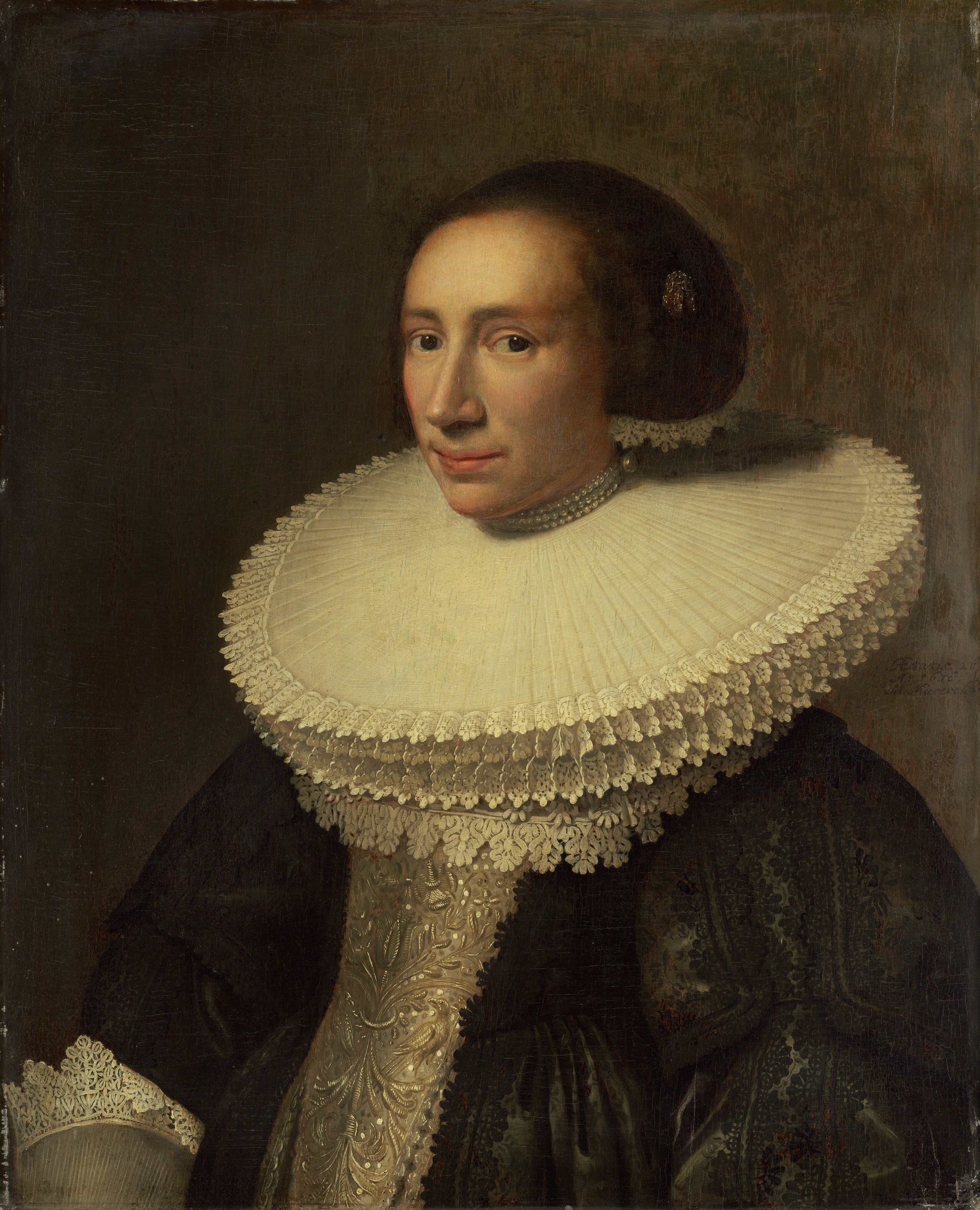 This painting shows a woman from the waist up, in a three-quarter view, facing the left. She has fair skin, a long nose, and narrow, pink lips. Her dark brown hair is styled in a large, smooth bun and accessorized with a golden brooch or pin and a strand of lace. Around her neck is an exaggeratedly large lace collar, with several layers of long, straight lace under three strands of pearls. She wears a dark gown with intricate patterns on the sleeves, a sliver bodice, and lace-trimmed cuffs. The background is plain gray.