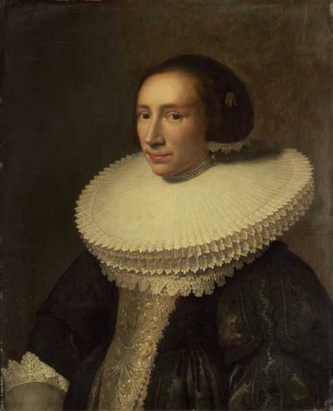 This painting shows a woman from the waist up, in a three-quarter view, facing the left. She has fair skin, a long nose, and narrow, pink lips. Her dark brown hair is styled in a large, smooth bun and accessorized with a golden brooch or pin and a strand of lace. Around her neck is an exaggeratedly large lace collar, with several layers of long, straight lace under three strands of pearls. She wears a dark gown with intricate patterns on the sleeves, a sliver bodice, and lace-trimmed cuffs. The background is plain gray.