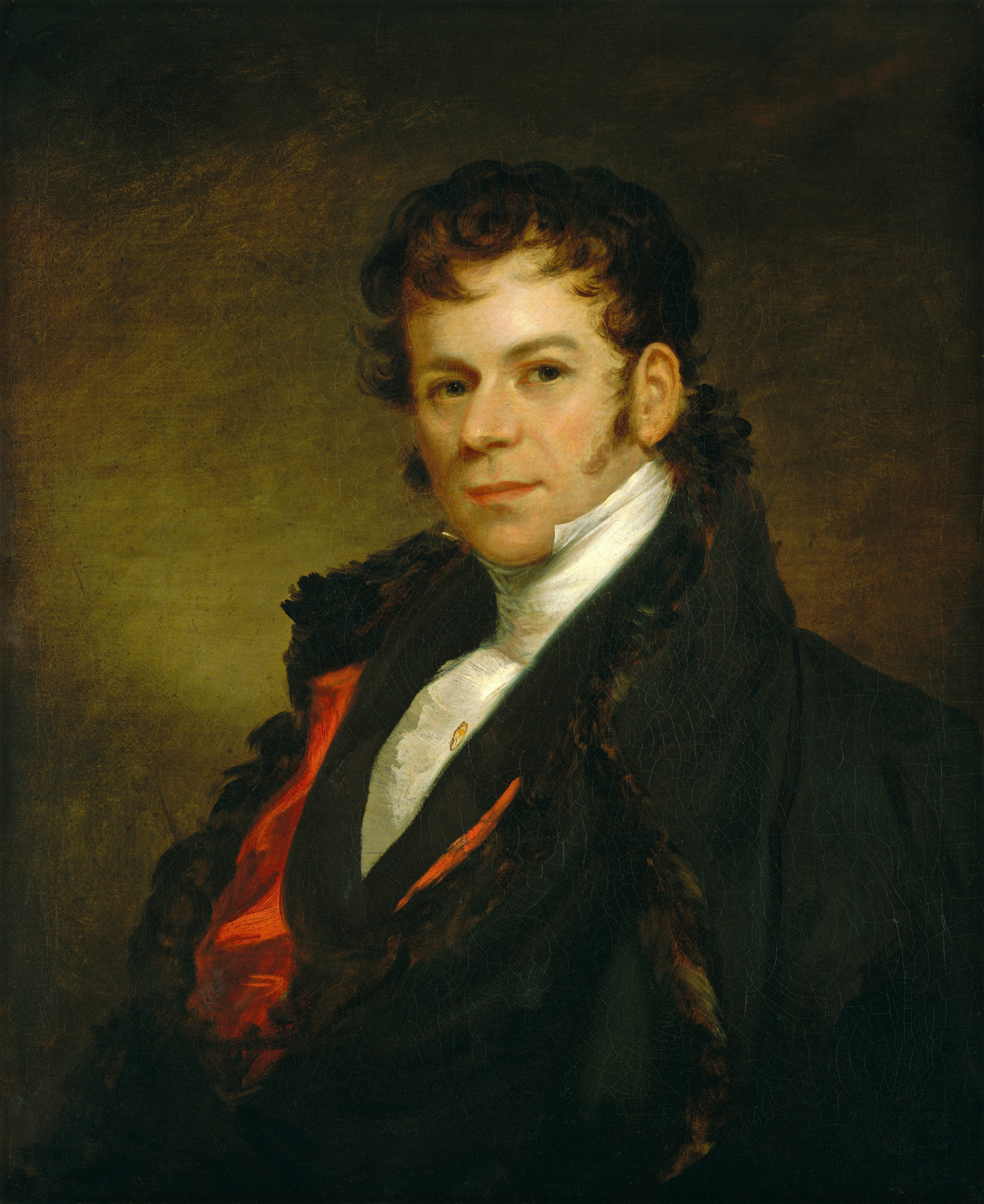 The image shows a man seated and facing forward. He has an angular face, sideburns, and dark, curly hair. His expression is neutral with a hint of a smile. He wears a dark coat with a fur-lined collar, a white high-collared shirt, and a red waistcoat. A pin decorates the waistcoat. The background is a dark, muted brown color.