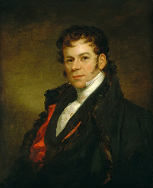 The image shows a man seated and facing forward. He has an angular face, sideburns, and dark, curly hair. His expression is neutral with a hint of a smile. He wears a dark coat with a fur-lined collar, a white high-collared shirt, and a red waistcoat. A pin decorates the waistcoat. The background is a dark, muted brown color.