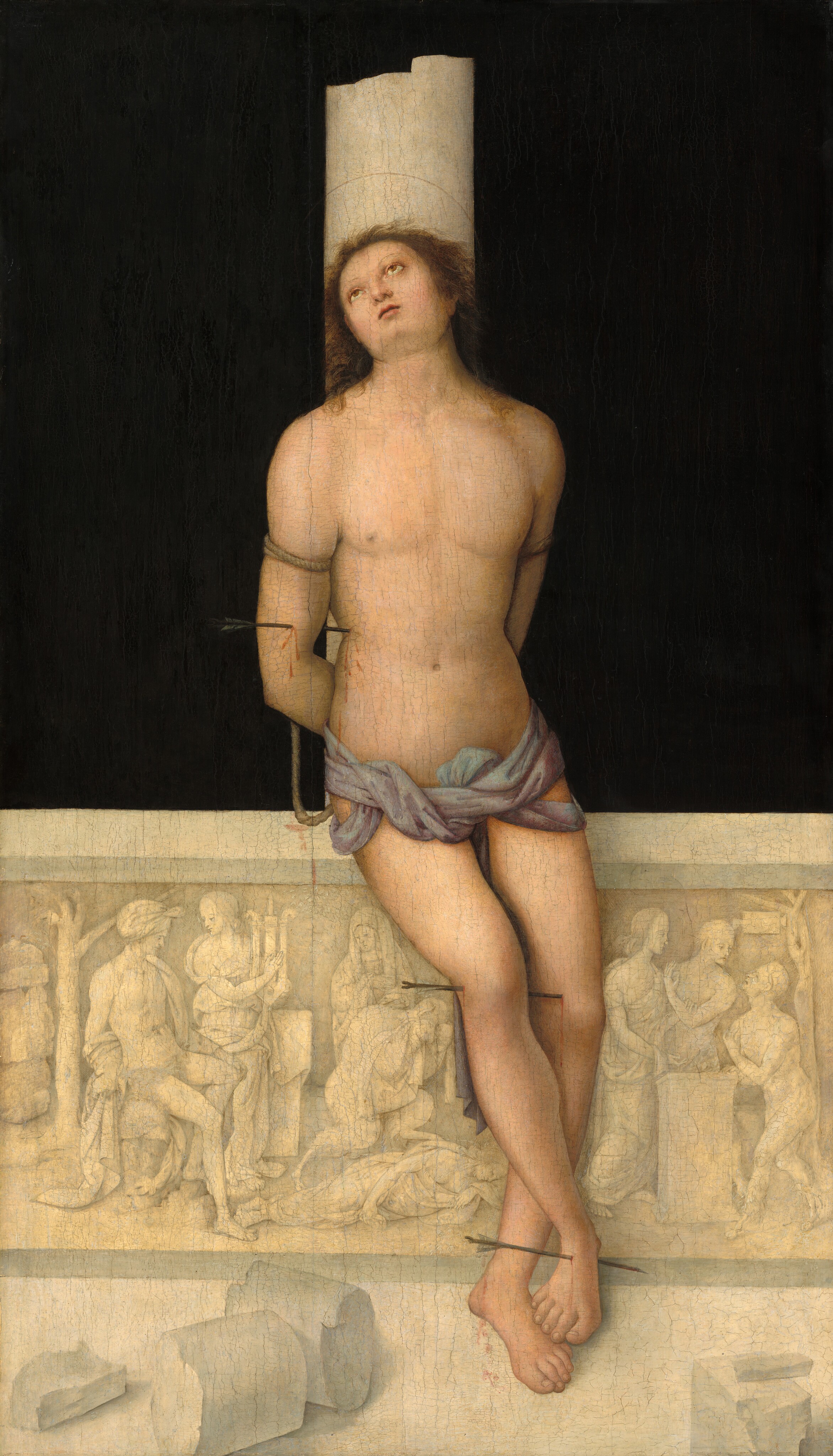 This painting shows a man leaning against a tall white column with his arms tied behind his back. He has light skin and shoulder-length brown hair, and his face is tilted upward with his eyes raised. He is pierced with arrows in several places on his body, including his arm, torso, knee, and foot. He is bare-chested and wears a draped, light purple cloth tied around his waist. The background is solid black, and the top of the column the man leans against is broken off. At the bottom of the column, there is a detailed white stone relief with figures in varying poses, along with some fragmented pieces of stone on the white stone ground.