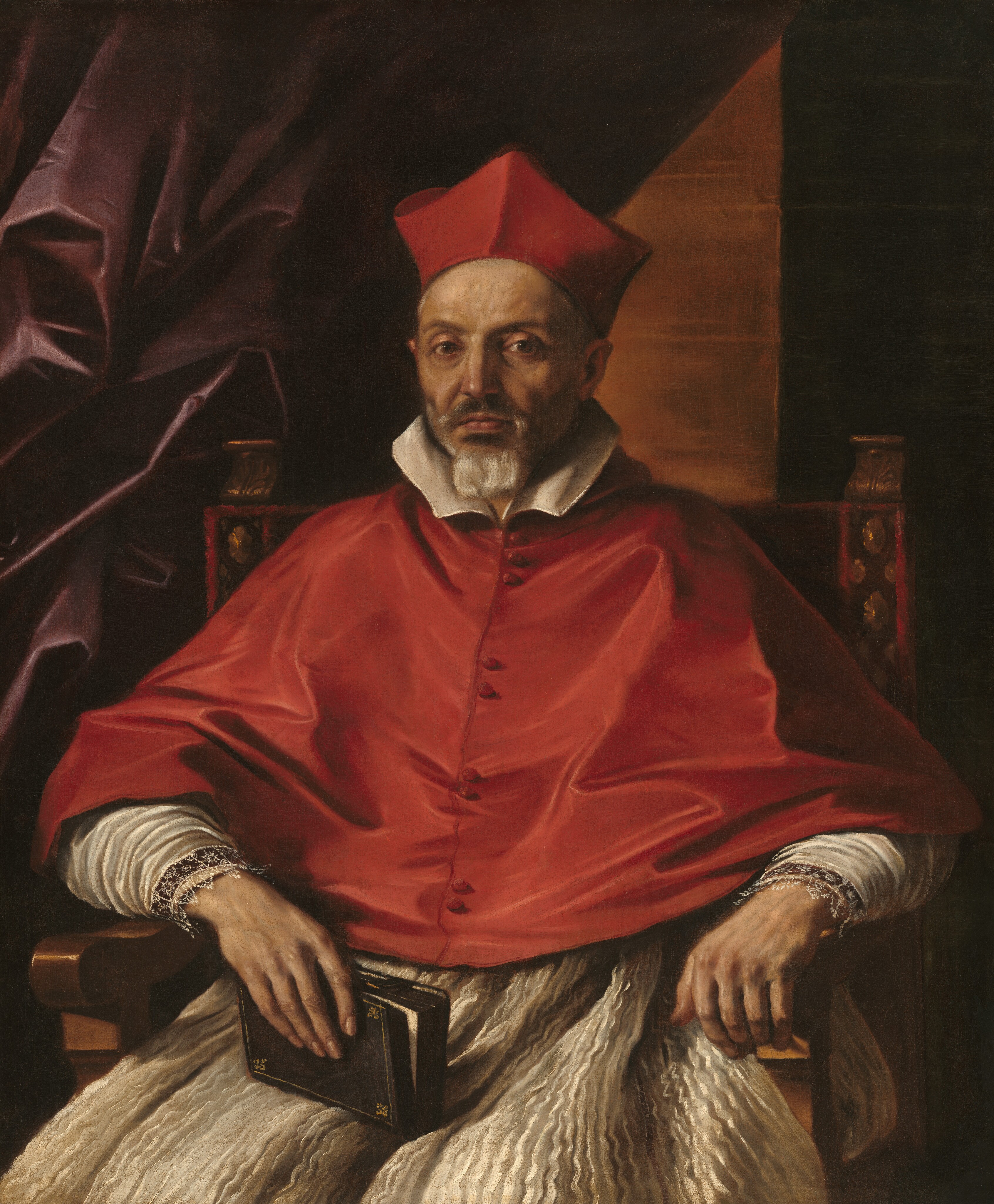 In this painting, a man is shown seated, facing forward, with a red biretta on his head. He has pale, wrinkled skin, deep-set eyes, a prominent nose, and a neatly trimmed grey beard and mustache. He wears a red garment over his shoulders and torso with a white collar and cuffs, and lace details on the sleeves, with a pleated white robe underneath. His forearms rest on the arms of the chair he sits in, and in one of his hands he holds a partially open book. The chair is ornately decorated, and the background shows dark purple drapery on the left and a dark wall on the right, with an illuminated triangle of lighter brown.